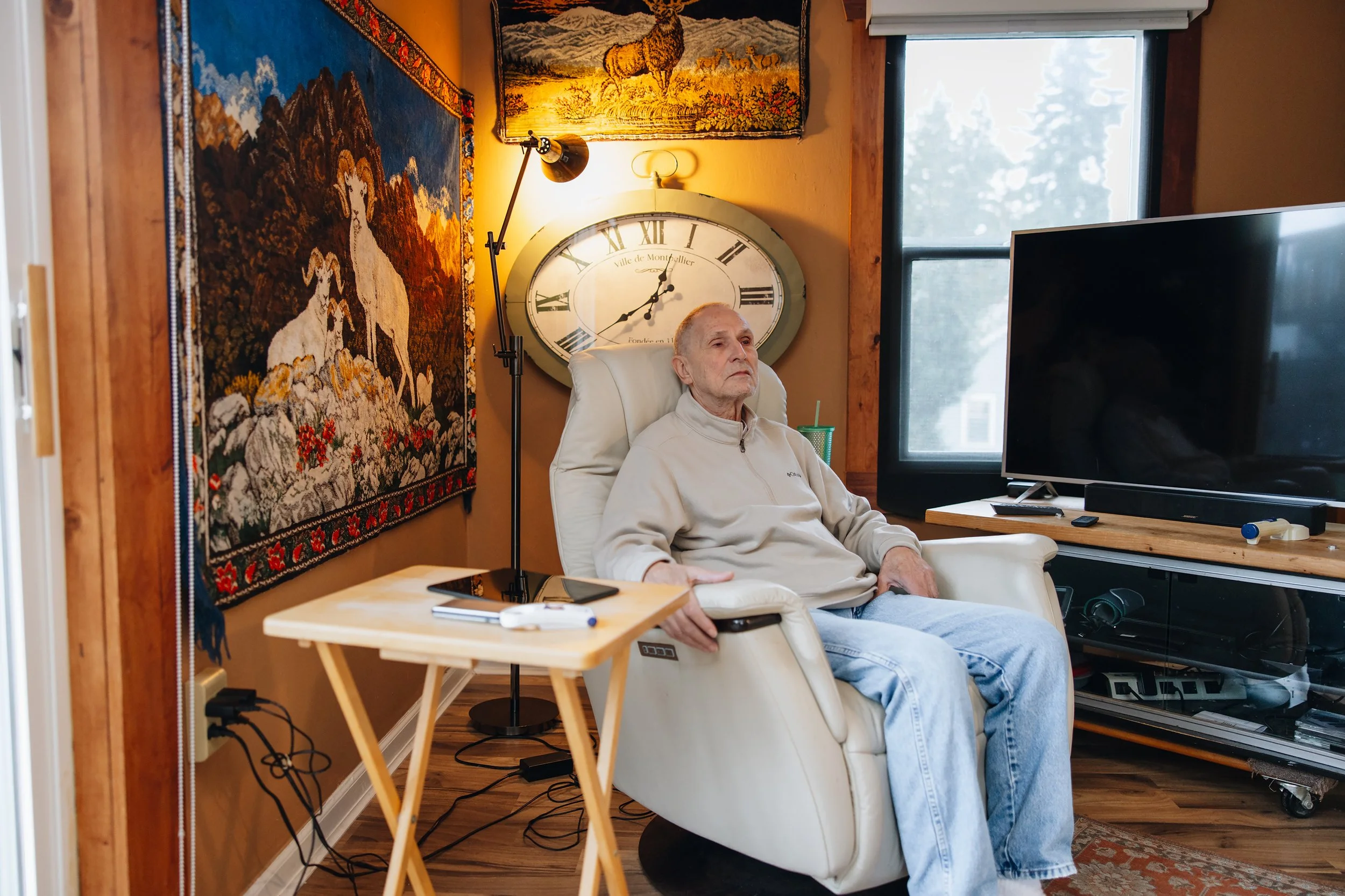An elderly man sitting in a white armchair in a cozy living room with wooden accents. Behind him is a large wall clock, and there are colorful tapestries on the wall featuring dogs and a landscape. To his right is a flat-screen TV on a wooden stand. On a small wooden table beside him are a smartphone and a drink with a straw. The room has a window with a view of trees outside.