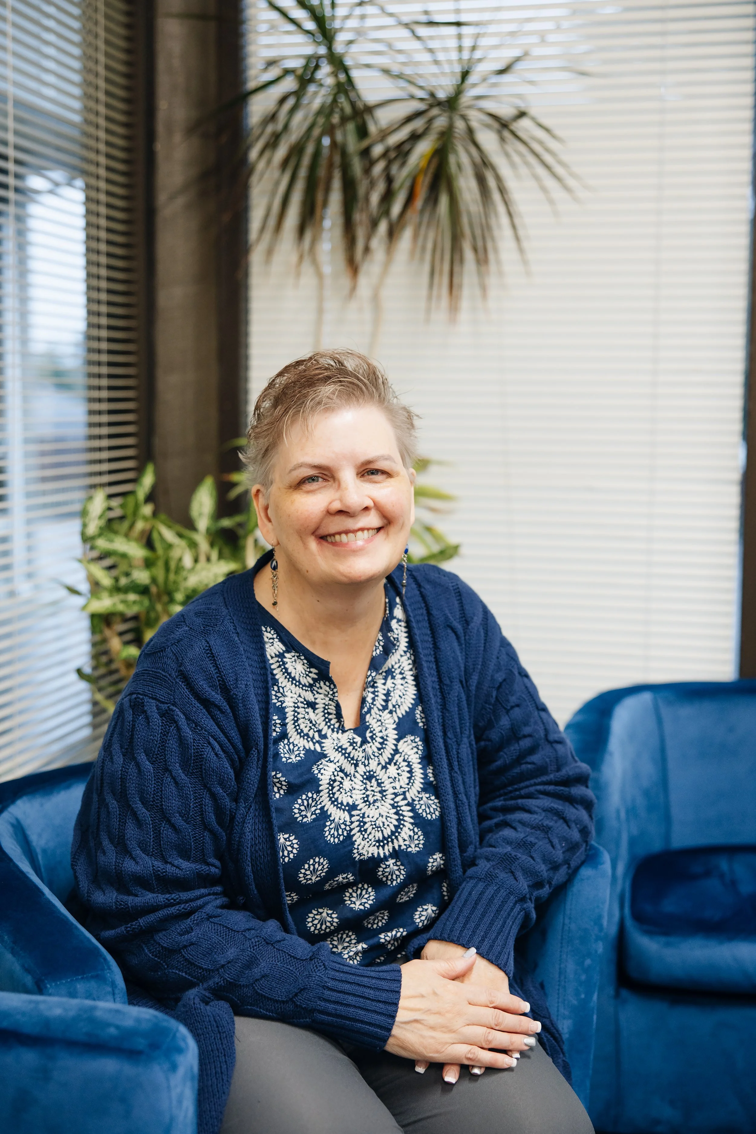 A smiling middle-aged woman with short gray hair, wearing a blue embroidered blouse and a navy blue cardigan, sitting on a blue velvet sofa in a well-lit room with plants and large windows with blinds.