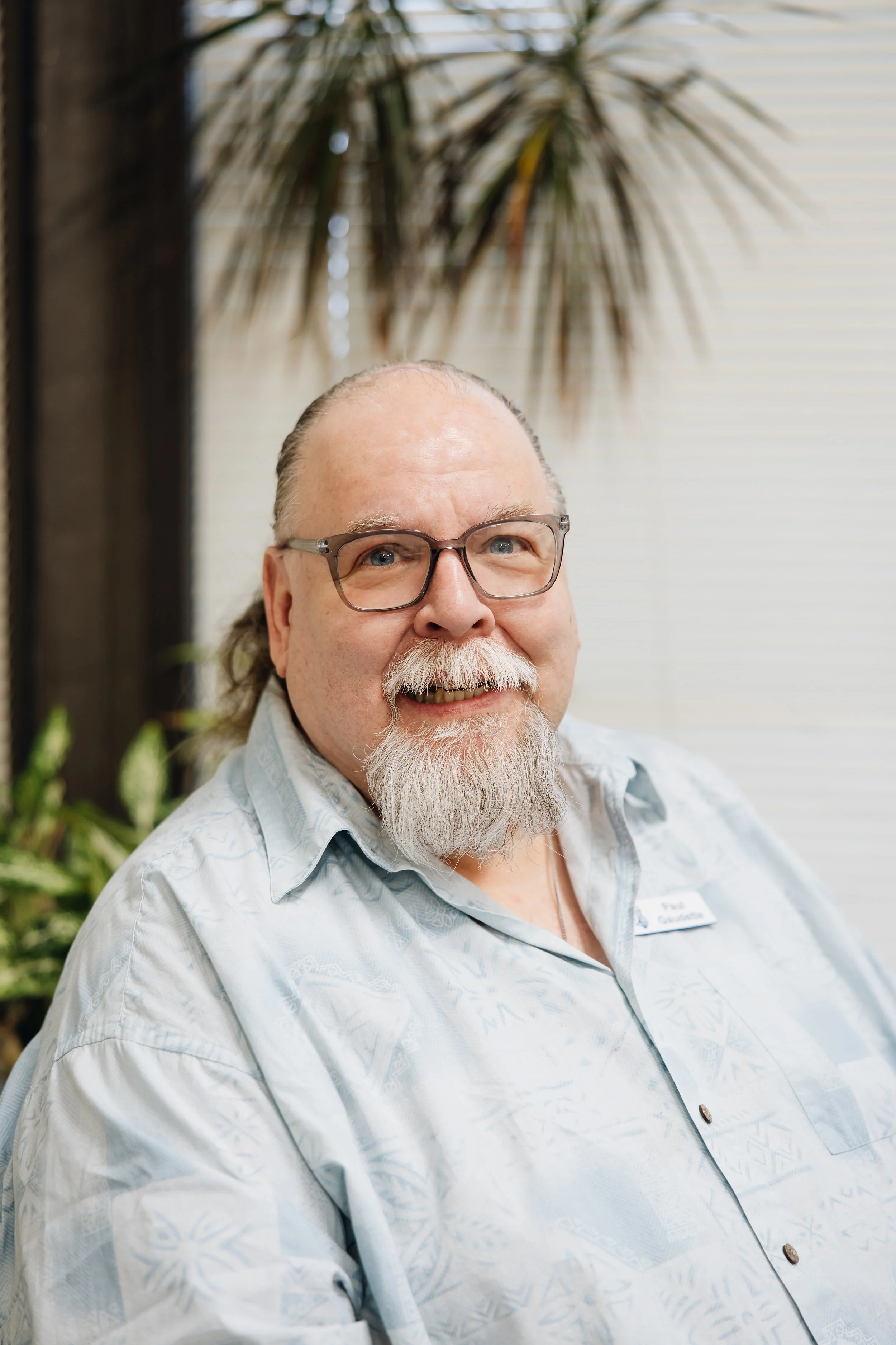 A smiling elderly man with glasses and a white beard, wearing a light-colored patterned shirt, sitting outdoors with a leafy plant and palm fronds in the background.