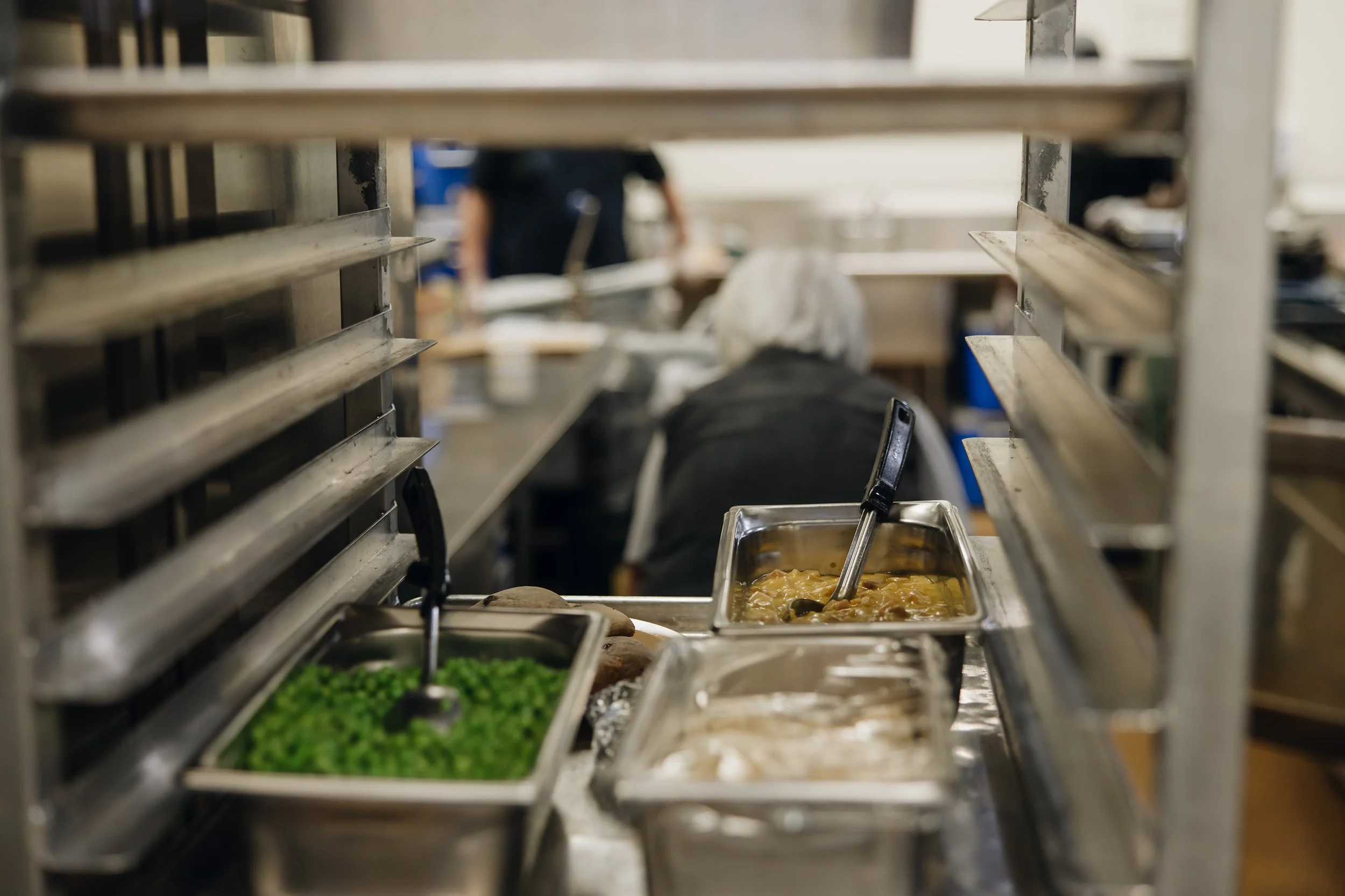 Food trays with peas and a stew in a cafeteria setting, viewed through metal shelves with people working in the background.