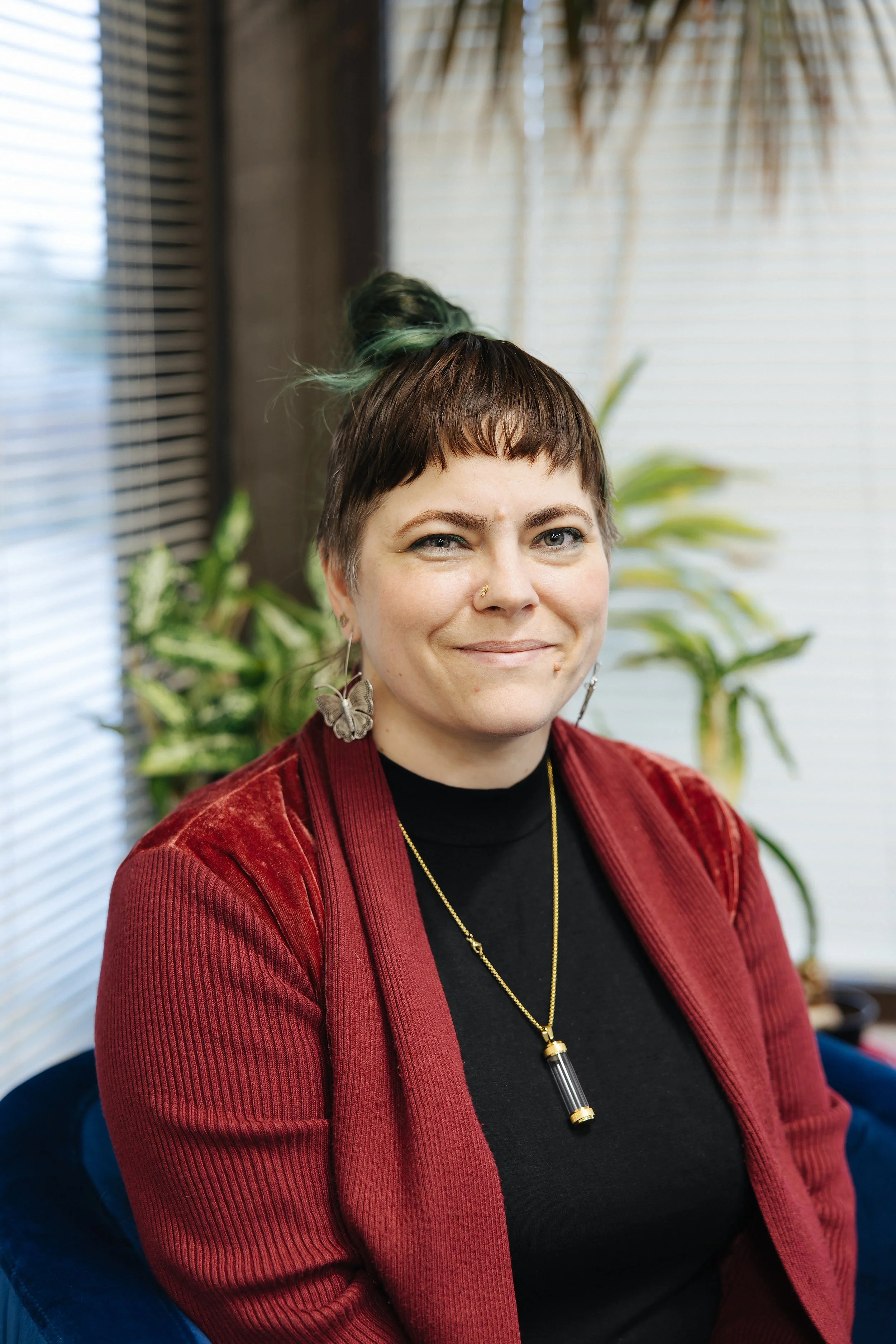 A woman with short hair, wearing a red velvet jacket, black shirt, and gold necklace with a pendant, sitting in front of a background with plants and window blinds.