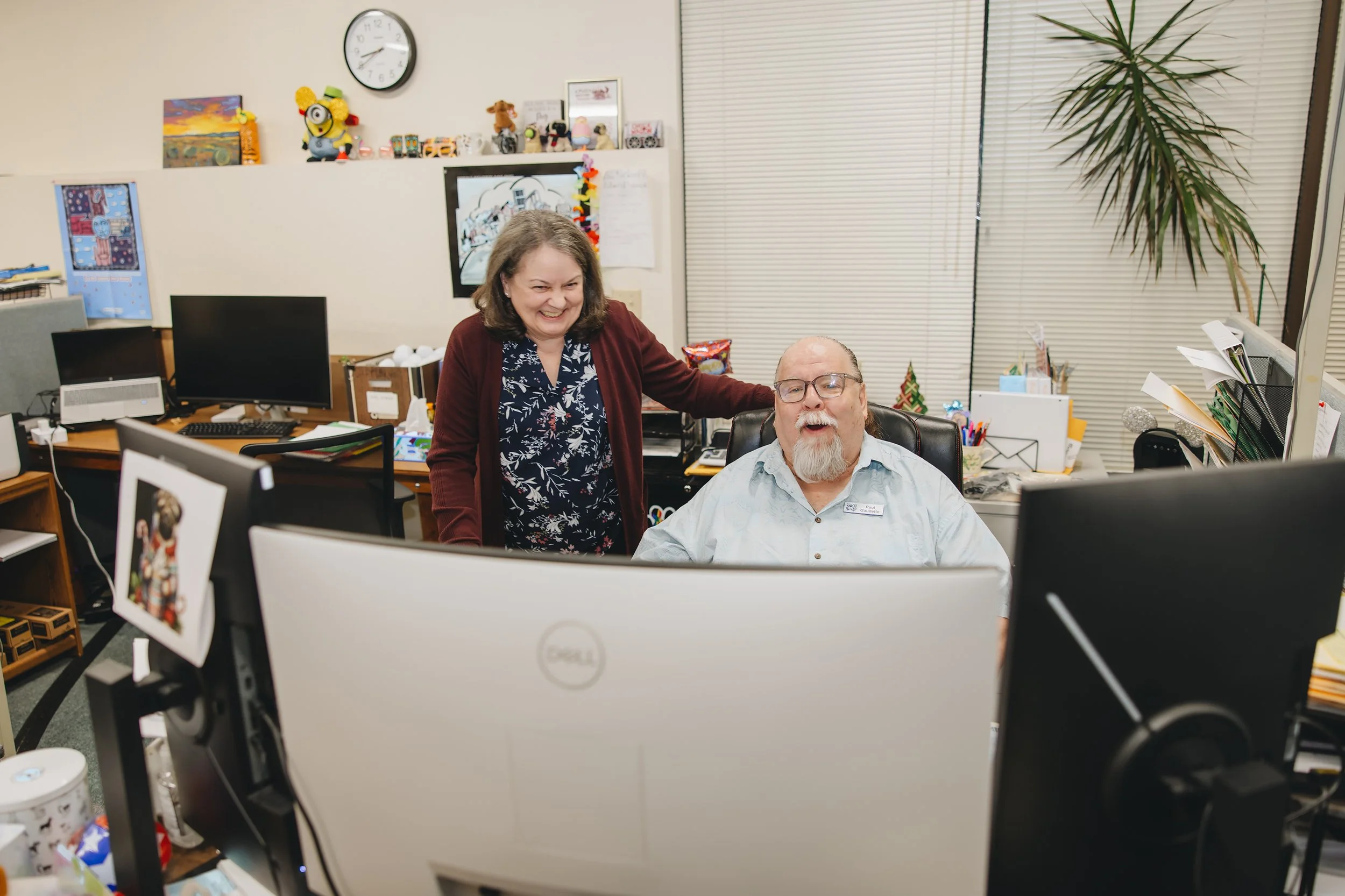 A woman standing next to a seated man in an office, both smiling and appearing happy, with computer monitors and office supplies in the background.