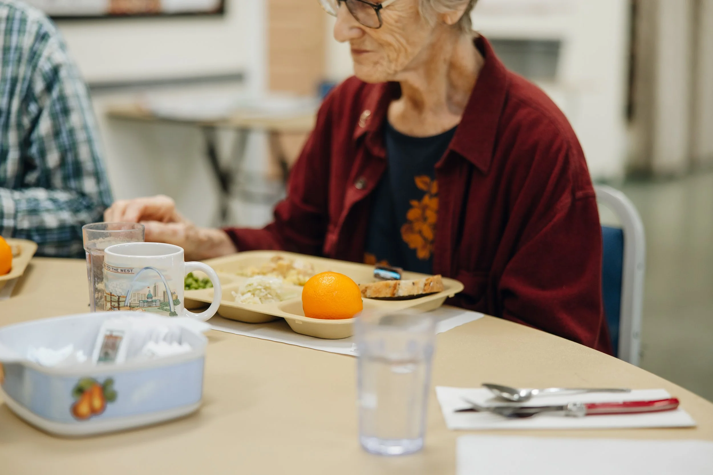 An elderly woman with glasses and a red shirt sitting at a dining table with a tray of food, an orange, and a mug, with other glasses and utensils on the table.