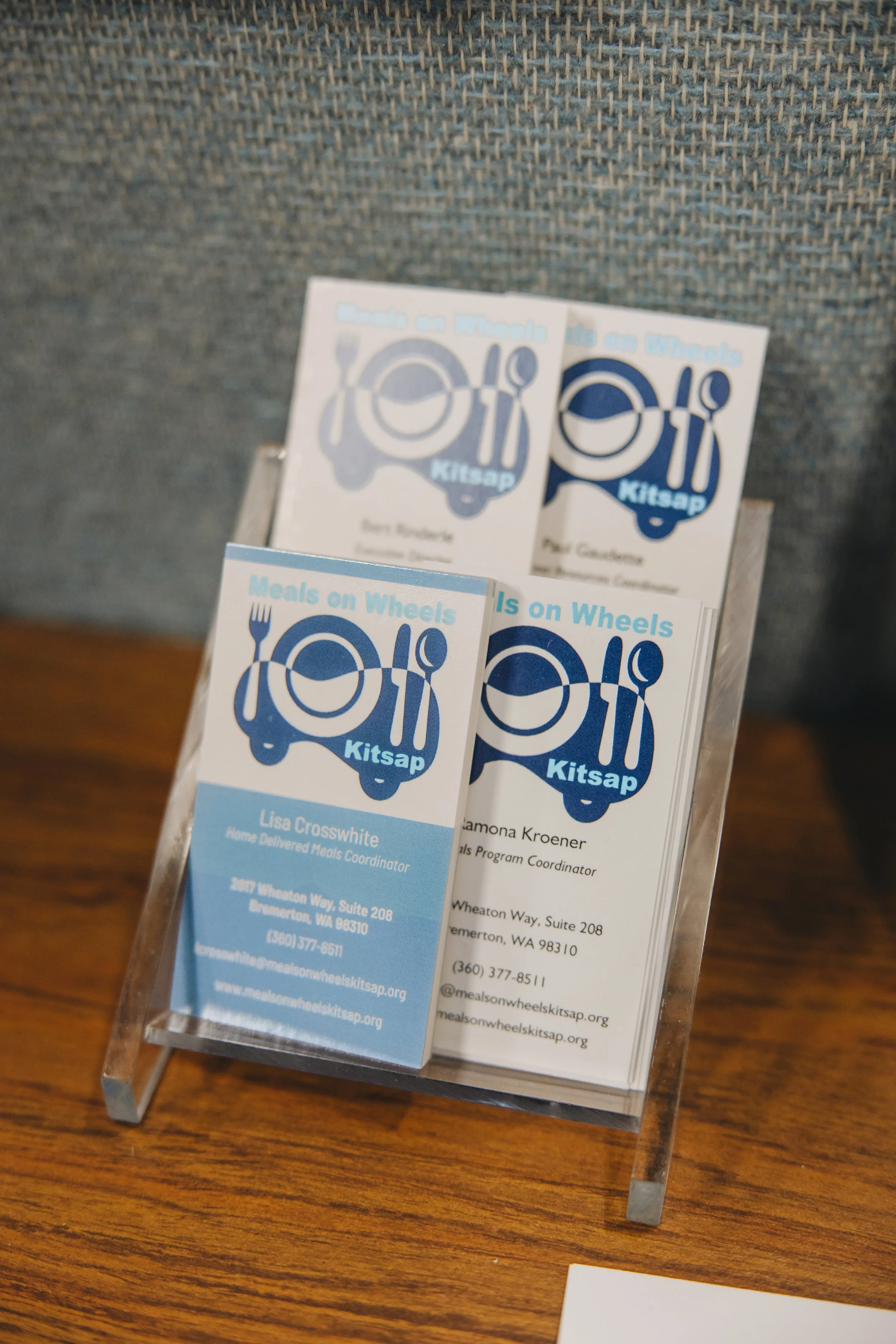 Business cards for Meals on Wheels Kitsap organization, displaying the logo, organization name, and contact information, placed in a clear acrylic holder on a wooden surface.
