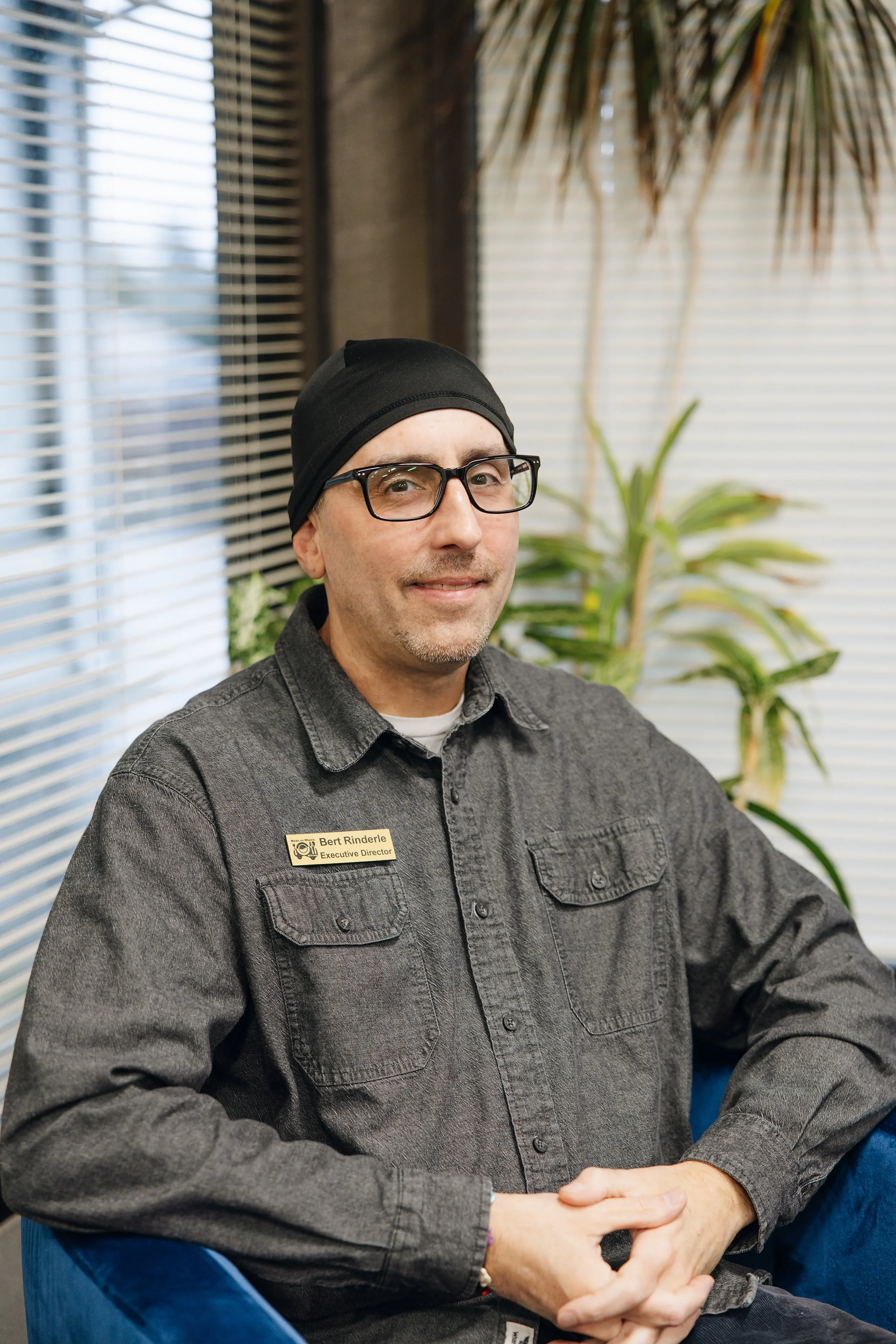 Man wearing black beanie, glasses, and a gray shirt with a nametag sitting indoors.