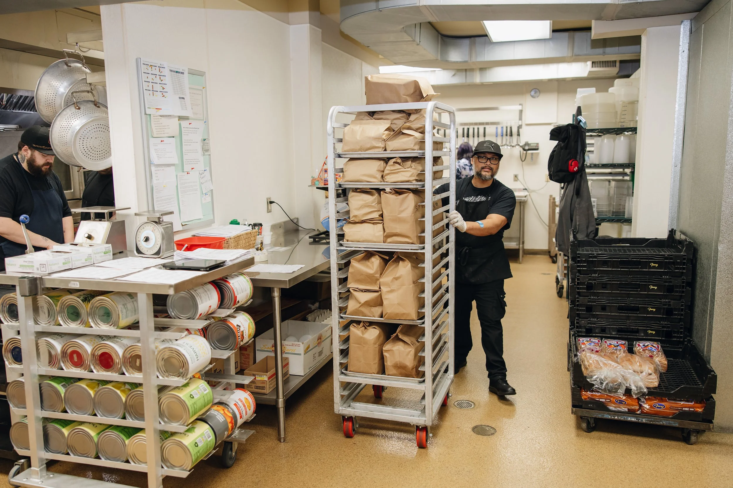 Two workers in a kitchen or food preparation area with carts of paper bags and bread, surrounded by canned goods and kitchen utensils.