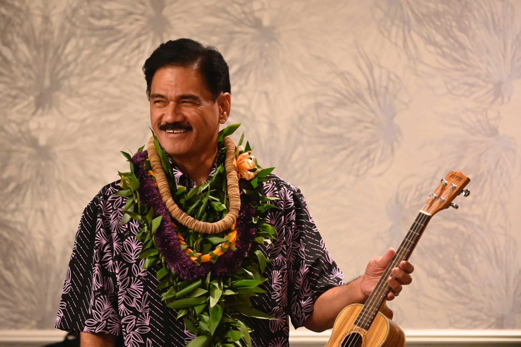 A man with a mustache wearing a floral patterned shirt, decorated with leis and a floral necklace, playing a ukulele.
