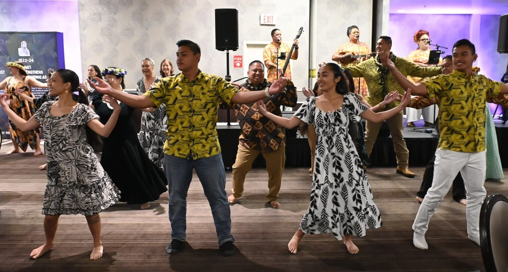 Group of people dancing barefoot in traditional Hawaiian attire with a band playing live music in the background at an indoor event.