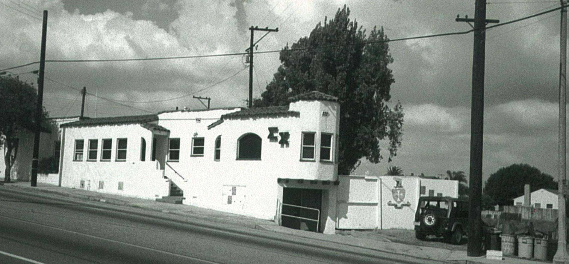 A black and white photo of a white stucco building with multiple windows, a staircase, and a decorative crest on the wall, located on a street with utility poles and a parked vehicle nearby.
