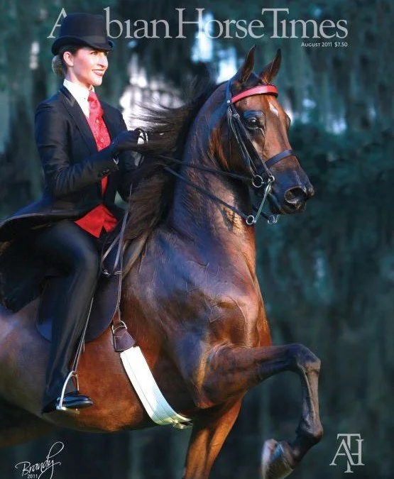 A woman riding a brown horse during daytime, both appear to be in motion, with the woman smiling and wearing formal riding attire, including a black jacket, red tie, and helmet, on the cover of 'Arabian Horse Times' magazine.