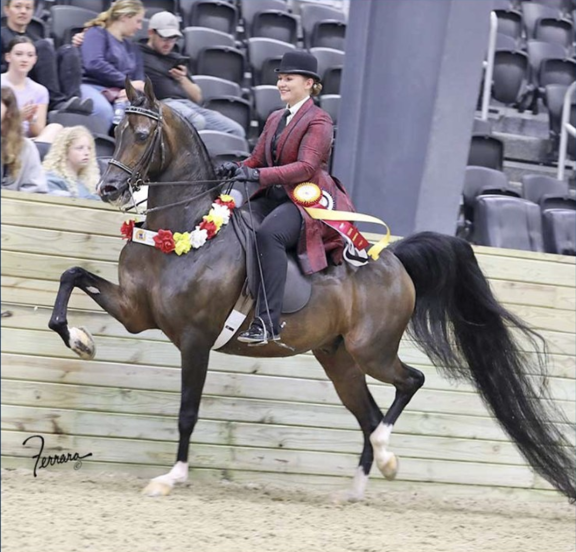 A woman in a red jacket, black helmet, and black riding gloves riding a brown horse with a flower garland decoration in an indoor equestrian arena. Spectators sit in the background watching.