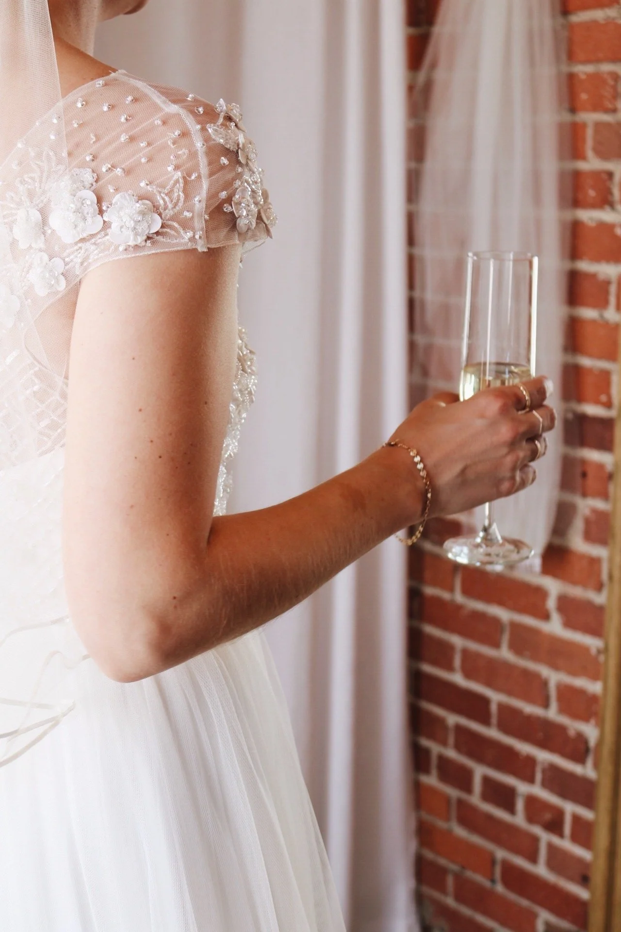 Close-up of a woman wearing a wedding dress with floral bead embellishments on the shoulder, holding a champagne flute, standing by a brick wall and curtain.