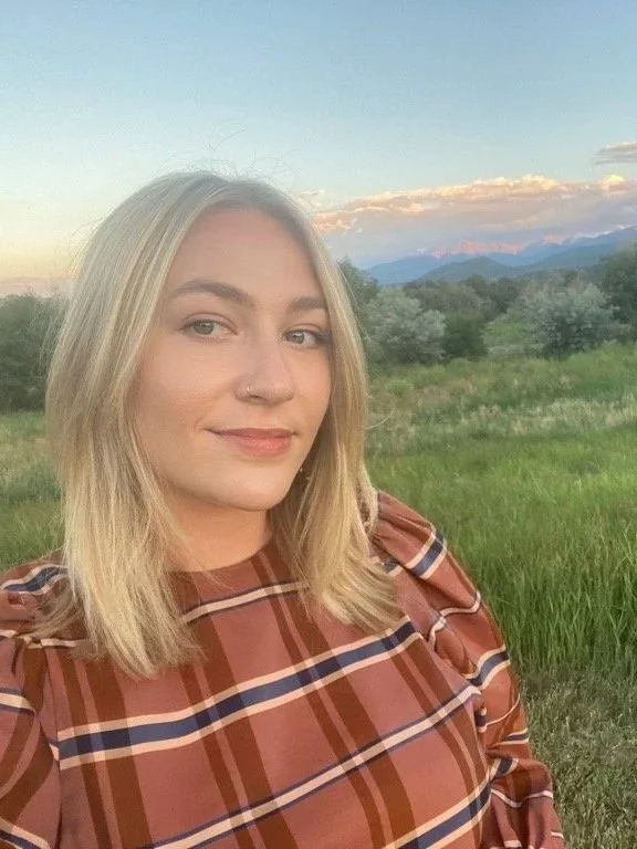 A young woman with blonde hair and a nose piercing taking a selfie outdoors during sunset, with mountains and greenery in the background.