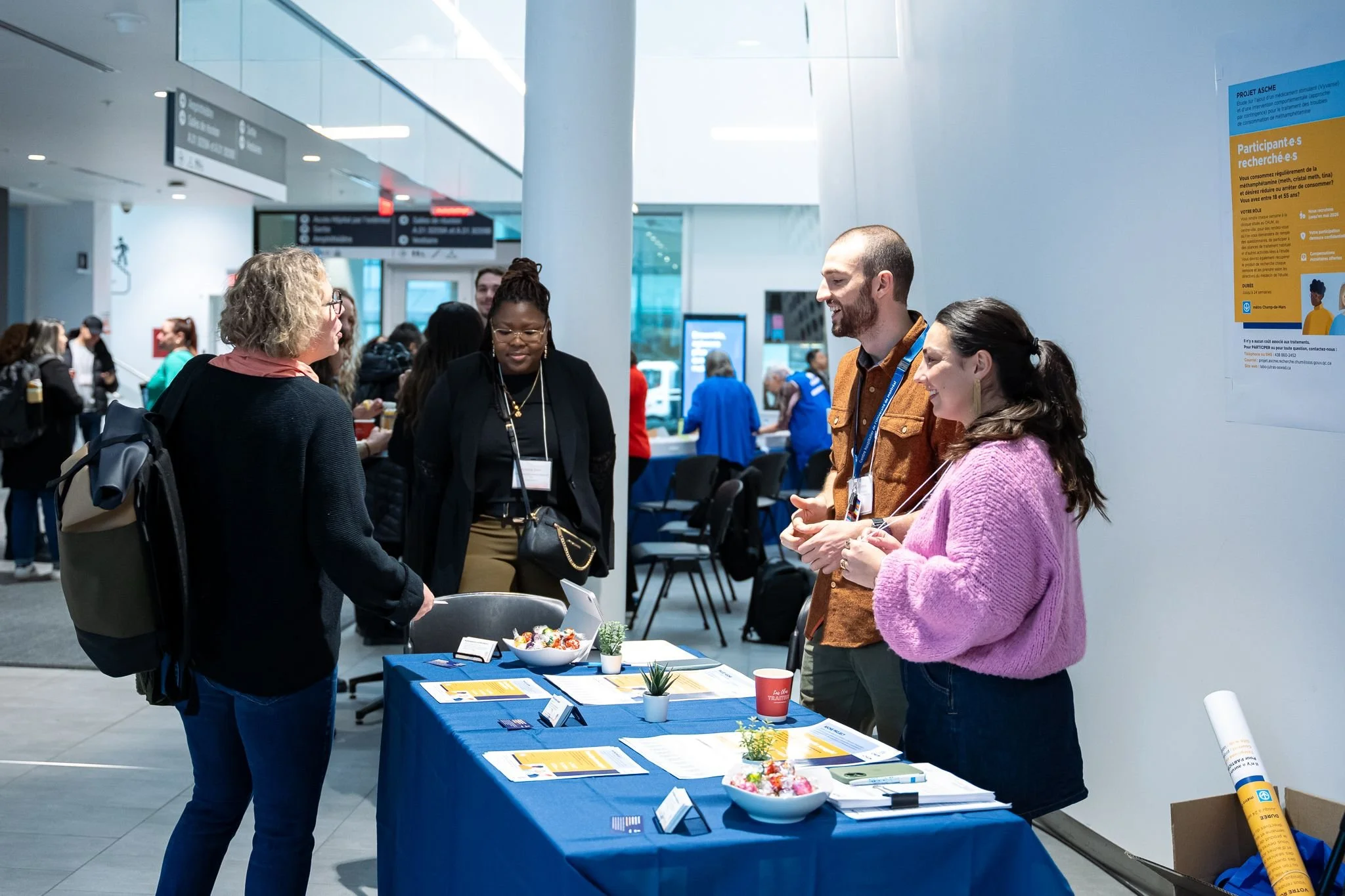 People at a conference registration or information booth, talking and exchanging information, with tables displaying pamphlets, flyers, and decorative bowls.