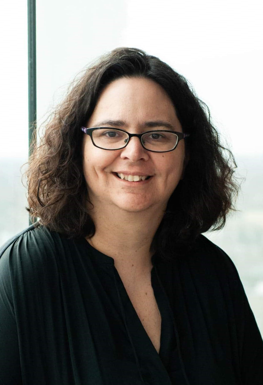 A smiling woman with curly dark hair and glasses, wearing a black top, standing near a large window with a view of the city skyline in the background.