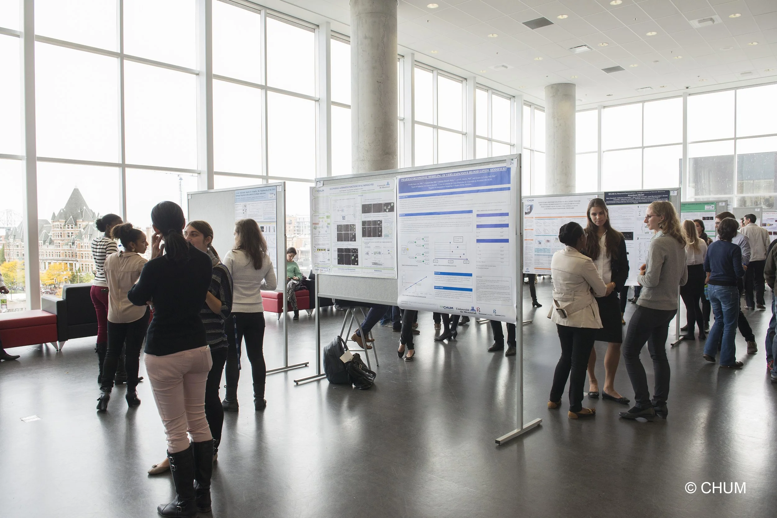 People attending a scientific conference or presentation in a modern building with large windows, displaying research posters on whiteboards, engaging in discussions.