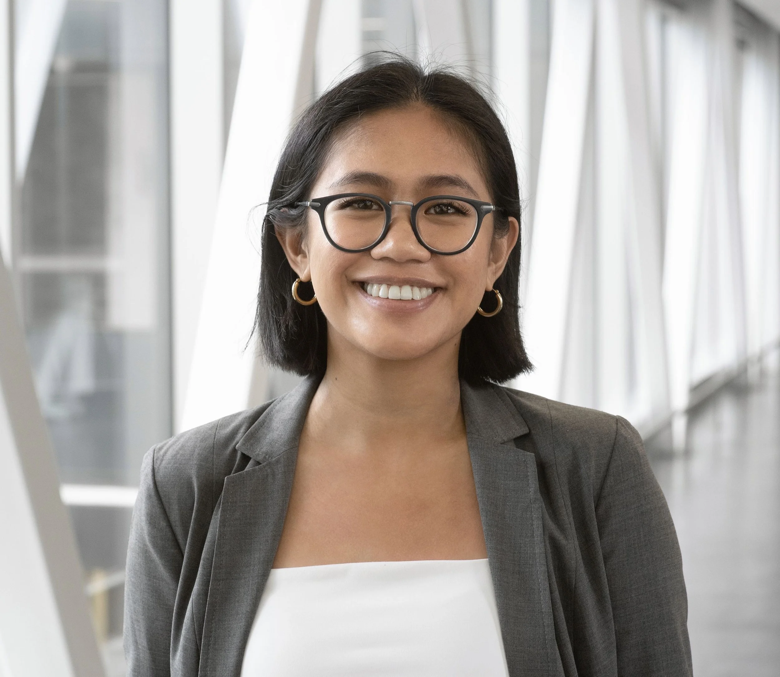 A smiling woman wearing glasses and earrings, dressed in a business suit, standing in an indoor space with bright lighting and modern architecture.