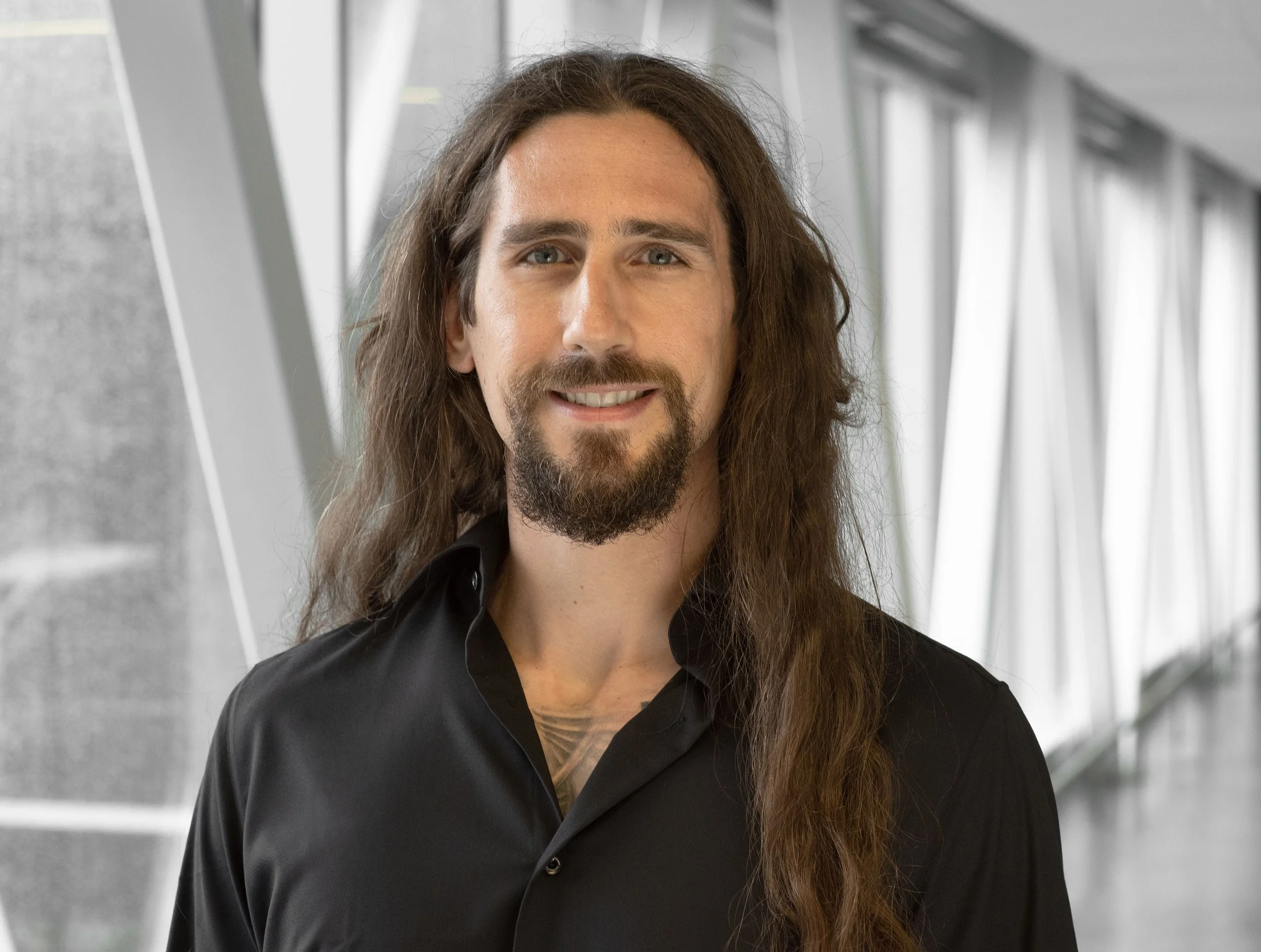 A man with long brown hair, a beard, and a mustache smiling, wearing a black shirt, in a modern building with large windows.