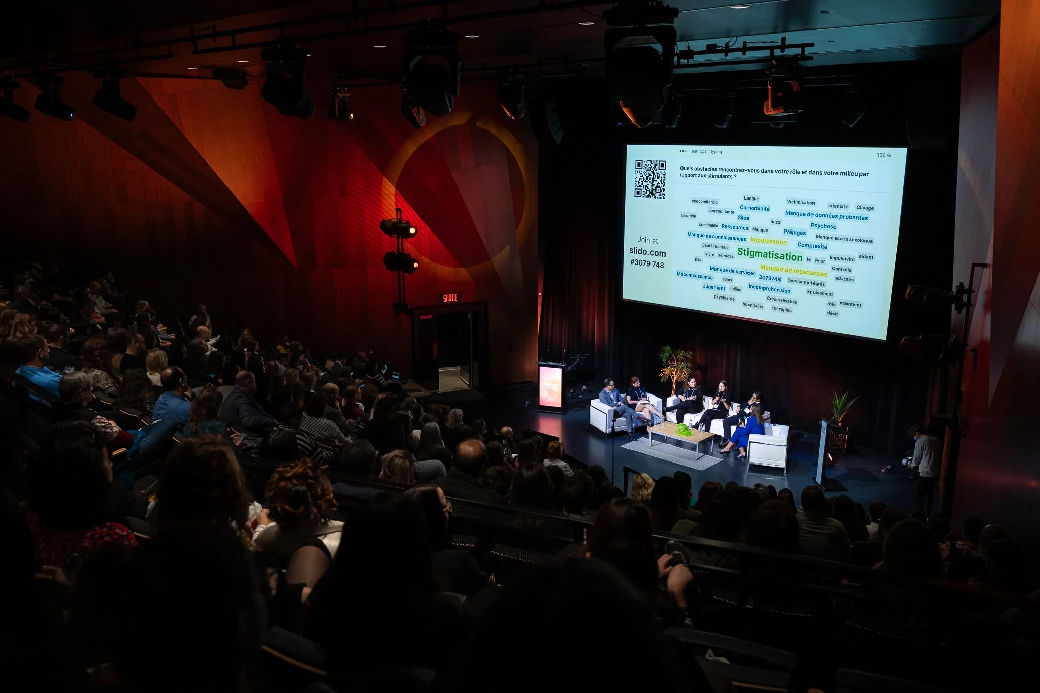 A large audience watching a panel discussion on a stage with a large screen displaying a quiz or presentation slide. The stage has five people seated, a table, and some plants. The screen shows text in French and a QR code.