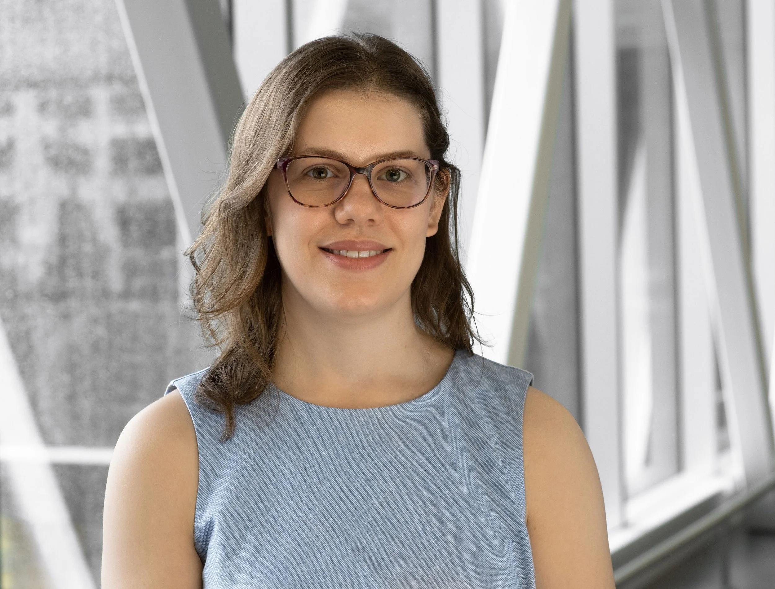 A woman with brown hair, wearing glasses and a sleeveless blue top, standing in front of a modern architectural background.