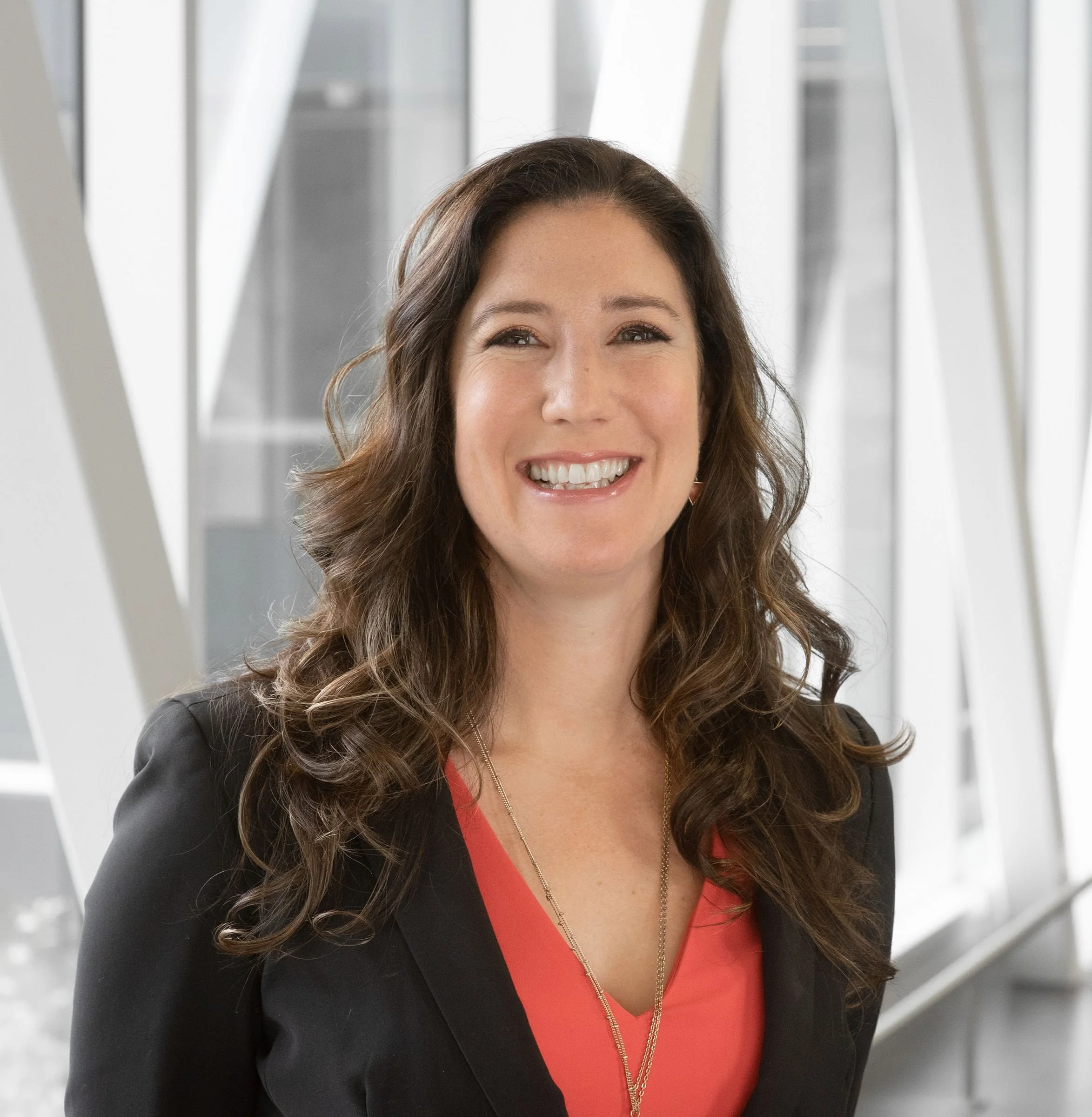 A woman with shoulder-length brown hair smiling, wearing a black blazer with a red top and gold necklace, standing in front of a modern architectural background.