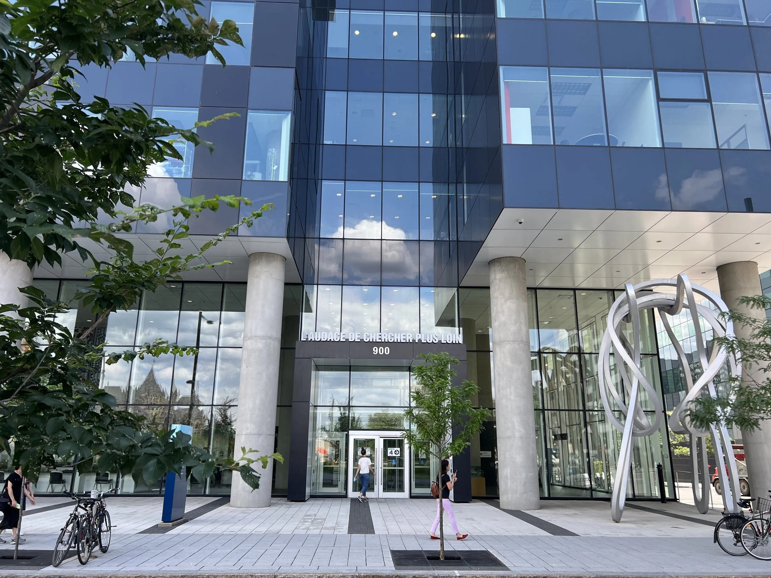 Modern glass building with the sign 'L'AUDACE DE CHERCHER PLUS LOIN' above the entrance, surrounded by trees, bicycles, and people walking outside.