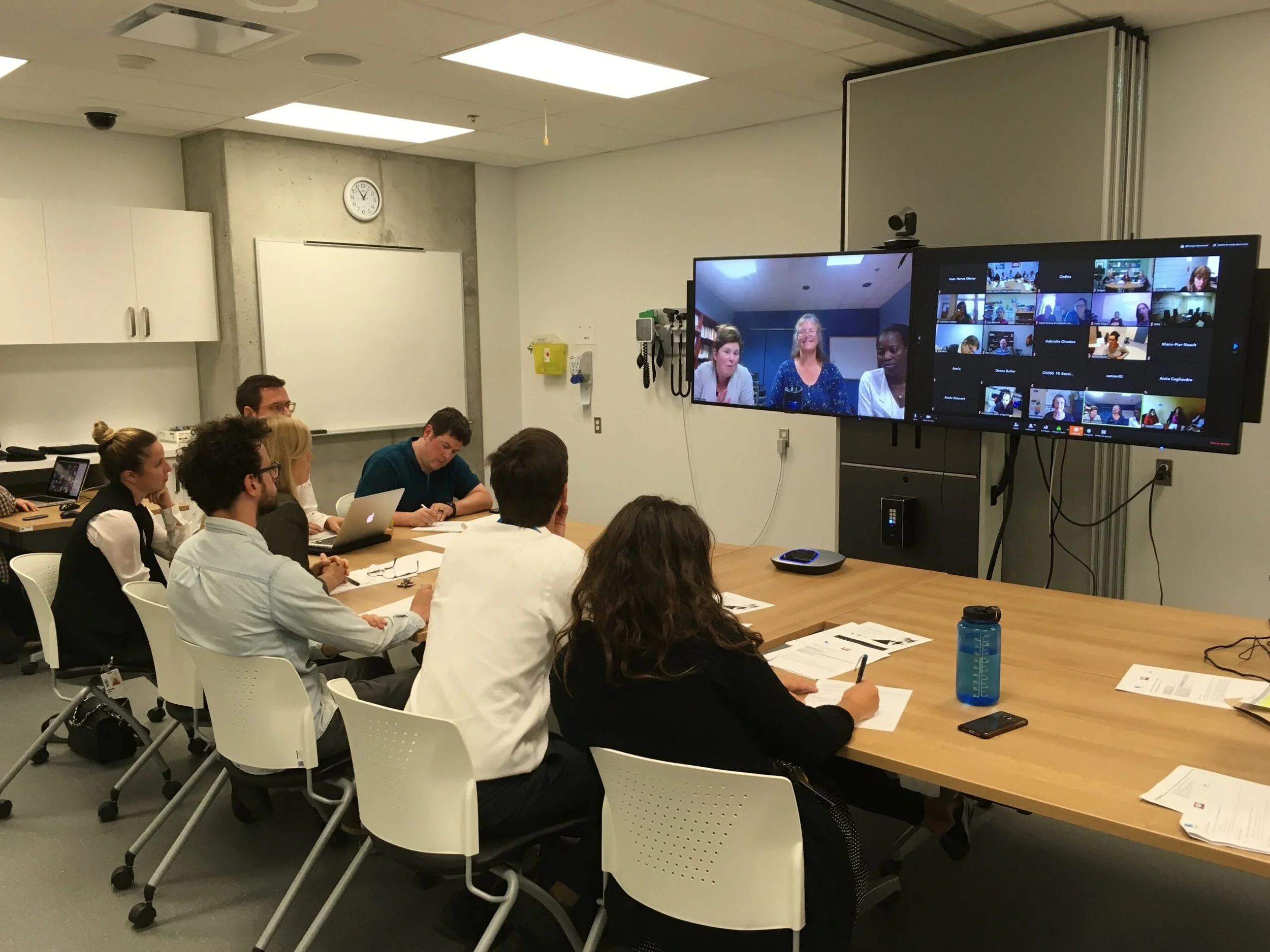 A group of people sitting around a conference table watching a video call on a large screen in a meeting room. The screen displays five women, while the room has a whiteboard, clock, and other office equipment.
