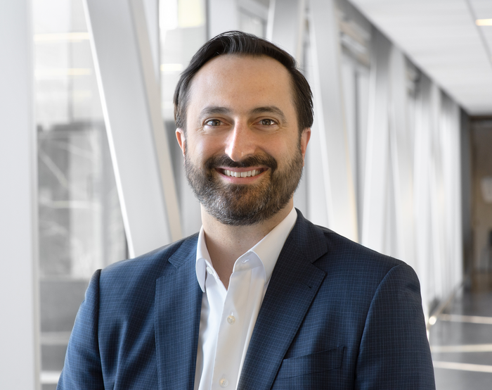 A man with dark hair and a beard wearing a navy blue suit and white shirt smiling in an office setting.