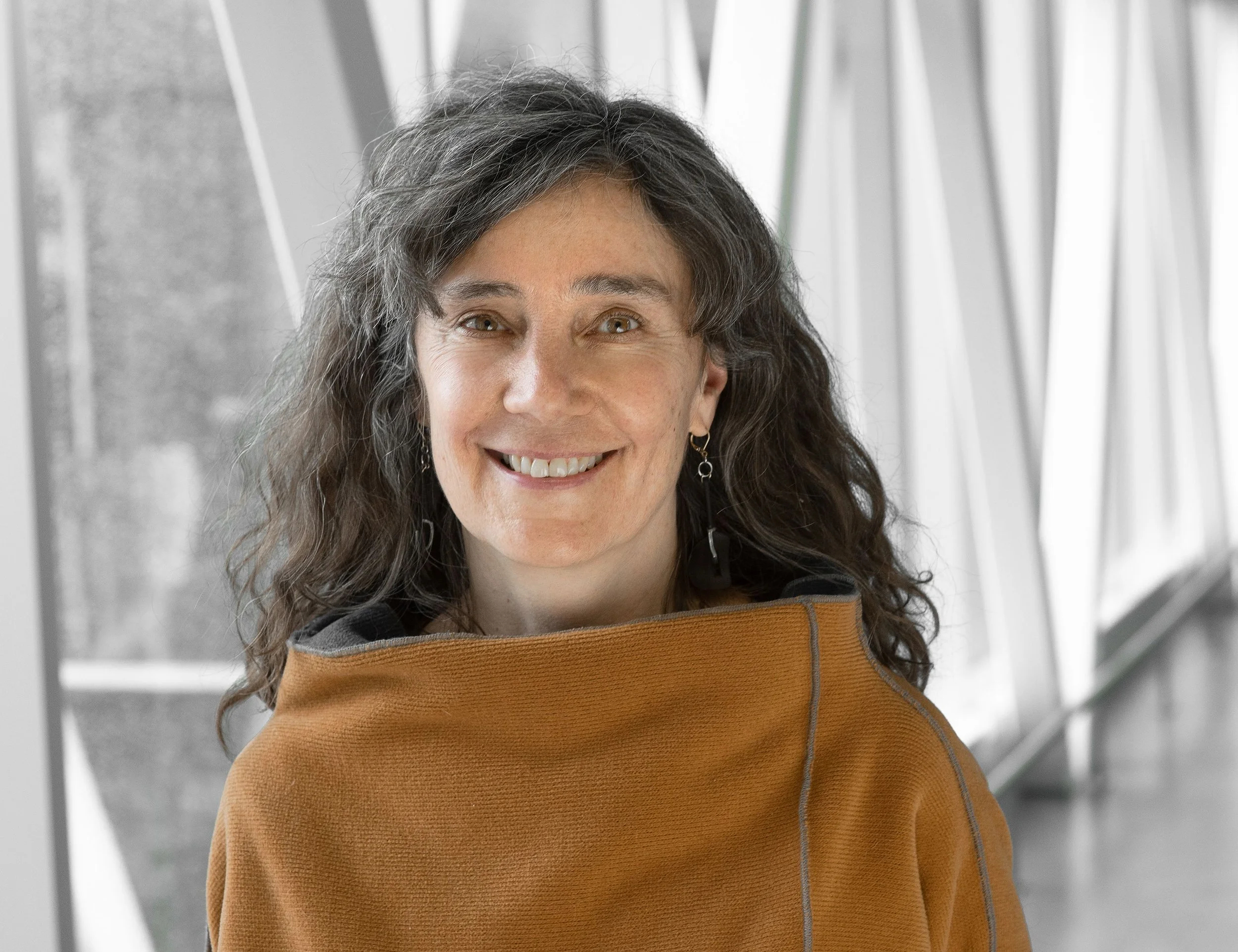 A woman with long, wavy dark hair smiling at the camera, wearing a brown sweater and earrings, with a background of metallic and glass structures.