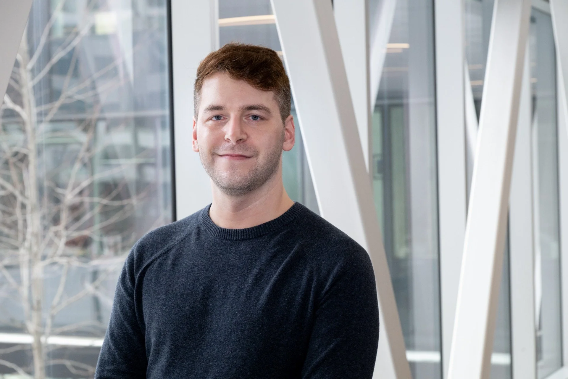 A young man with short brown hair and light skin, wearing a black sweater, standing indoors near large windows with a modern architectural background.