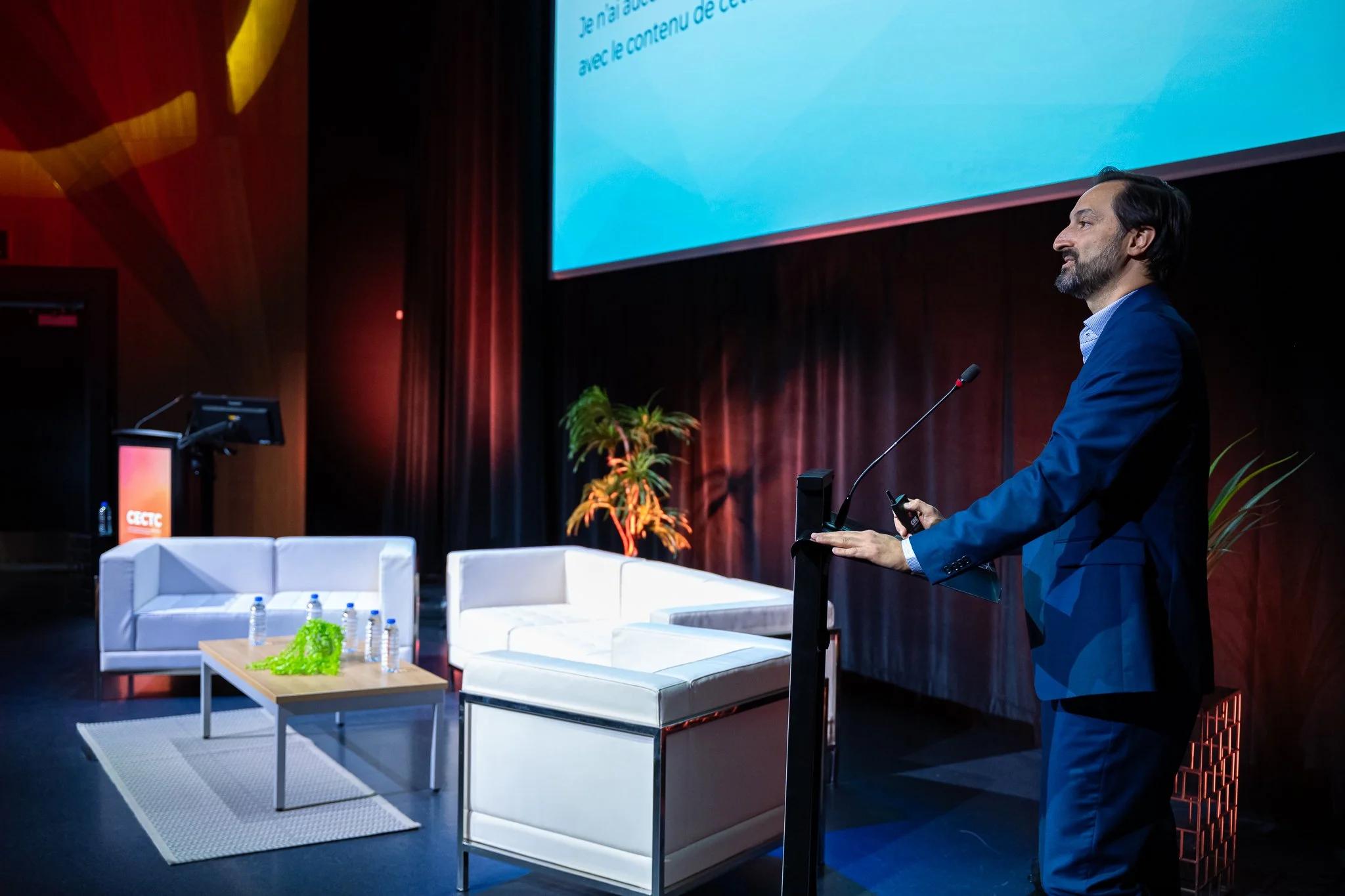 A man in a blue suit standing at a podium on stage during a conference or presentation. The stage has white chairs, a table with water bottles, and potted plants. A large screen display behind him shows text. The background includes wooden paneling and a dark curtain.