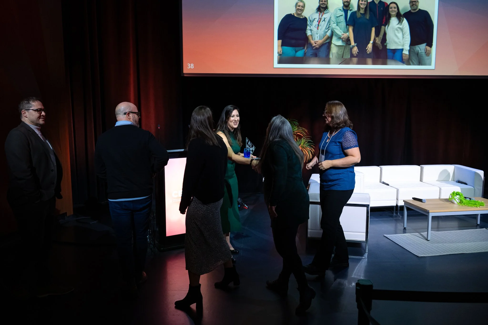 Group of five women and three men standing and conversing on stage at a conference or event, with a presentation slide projected behind them showing a group photo of seven people.