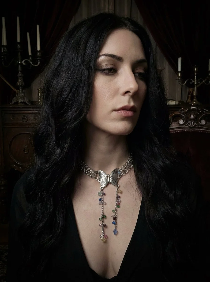 A woman with long black hair wearing a butterfly-shaped necklace with colorful dangling charms, dressed in black, in a dimly lit room with antique furniture and candle holders.