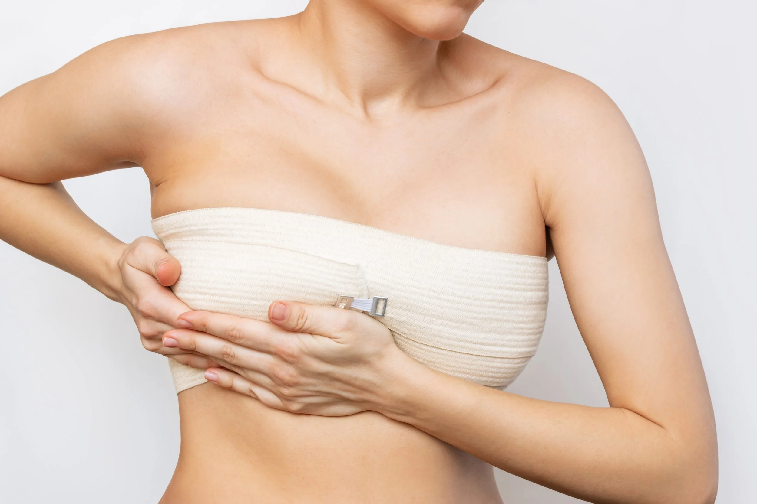 A woman wearing a strapless beige top, holding her left breast with her right hand, after breast augmentation surgery against a plain white background.