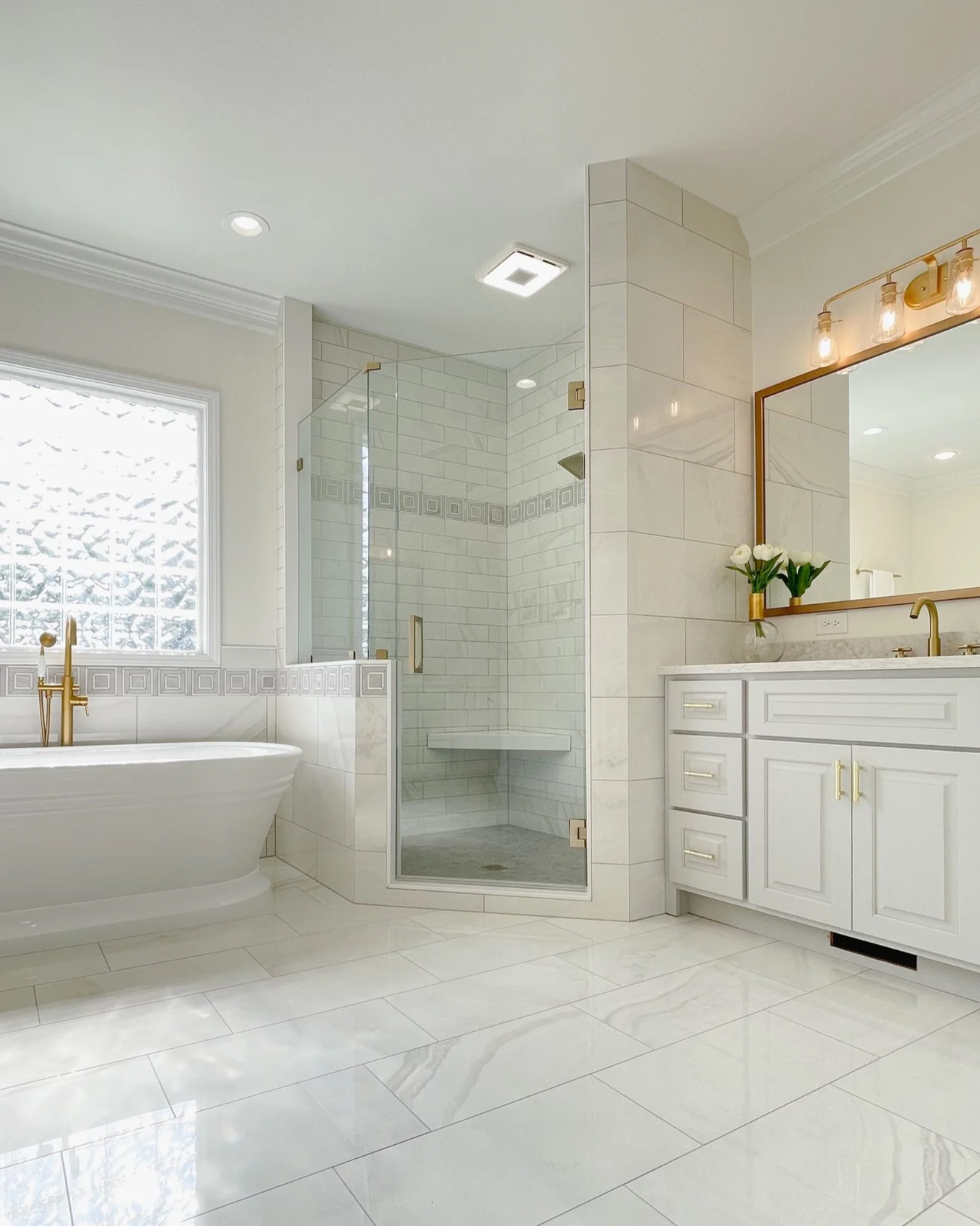 Bright modern bathroom with a white bathtub, glass-enclosed shower, white cabinetry with gold hardware, marble countertop, large mirror, and fresh flowers in a vase.