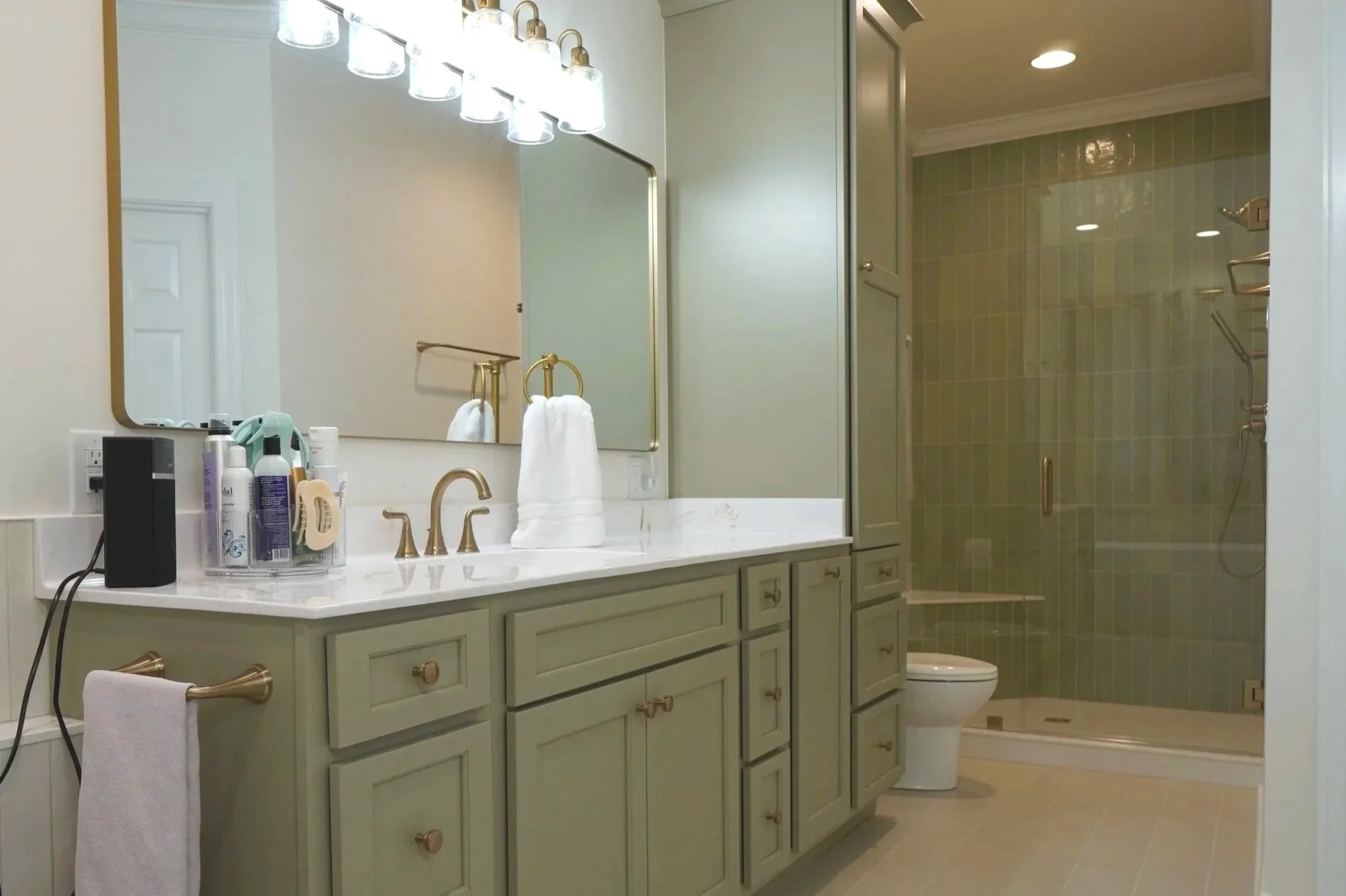 A bathroom with a large mirror above a white marble counter with gold fixtures. Various toiletries are on the counter. Towels hang on gold towel rings. There is a vanity cabinet below the counter. In the back, a glass-enclosed shower area with green tiles and gold fixtures, with a bathroom toilet nearby.