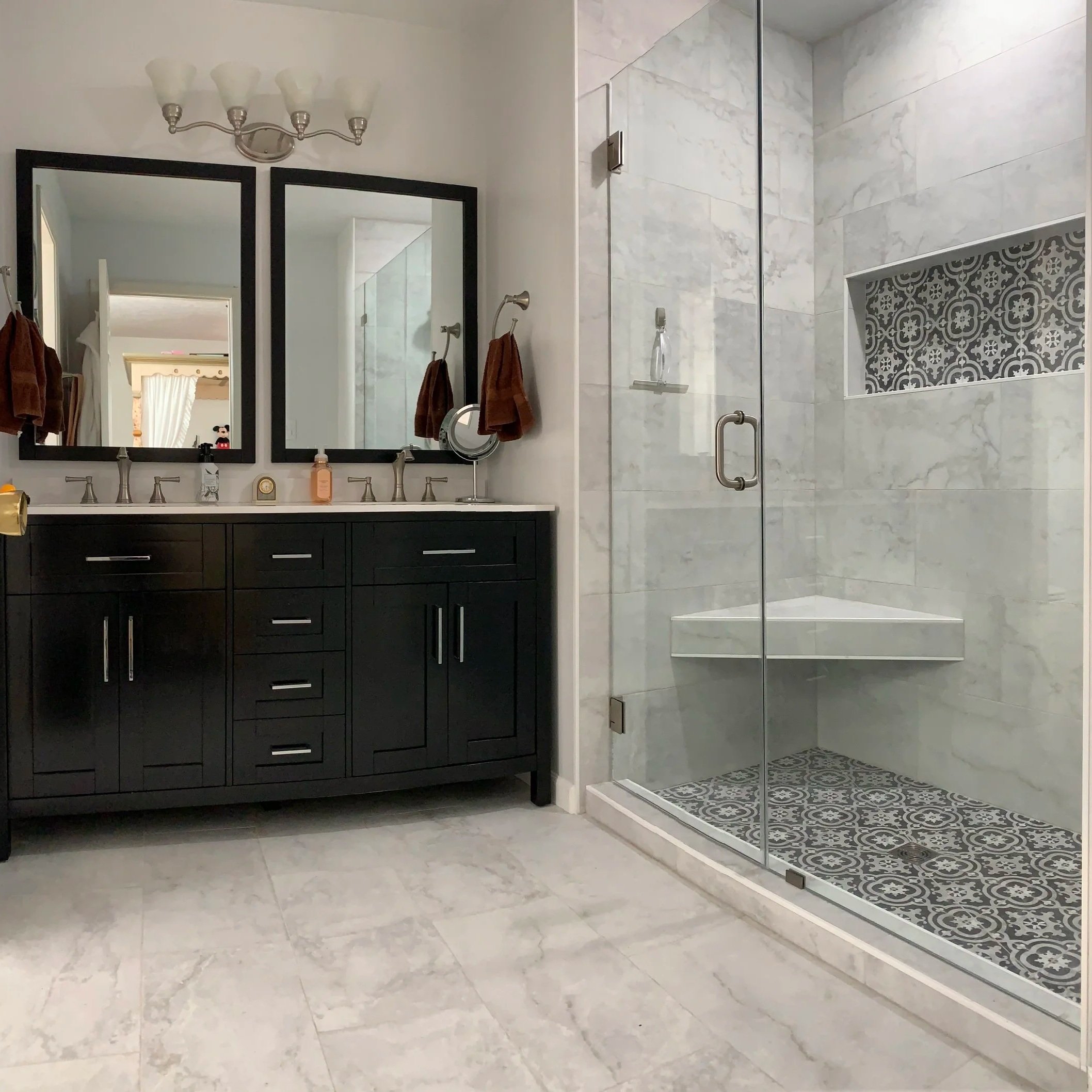 A modern bathroom with a black double vanity, two mirrors, brown towels, and a glass shower enclosure with decorative tile accents and a built-in bench.