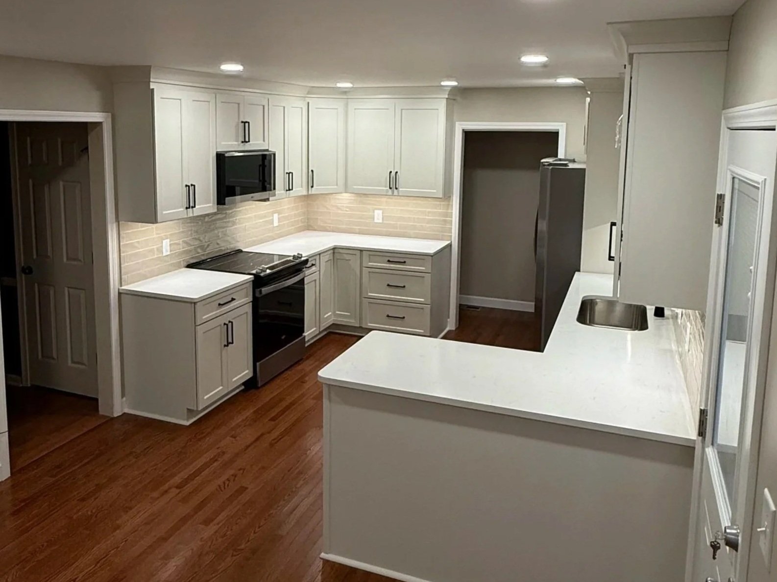 Modern kitchen with white cabinets, black appliances including microwave and stove, beige brick backsplash, white countertops, and hardwood flooring.