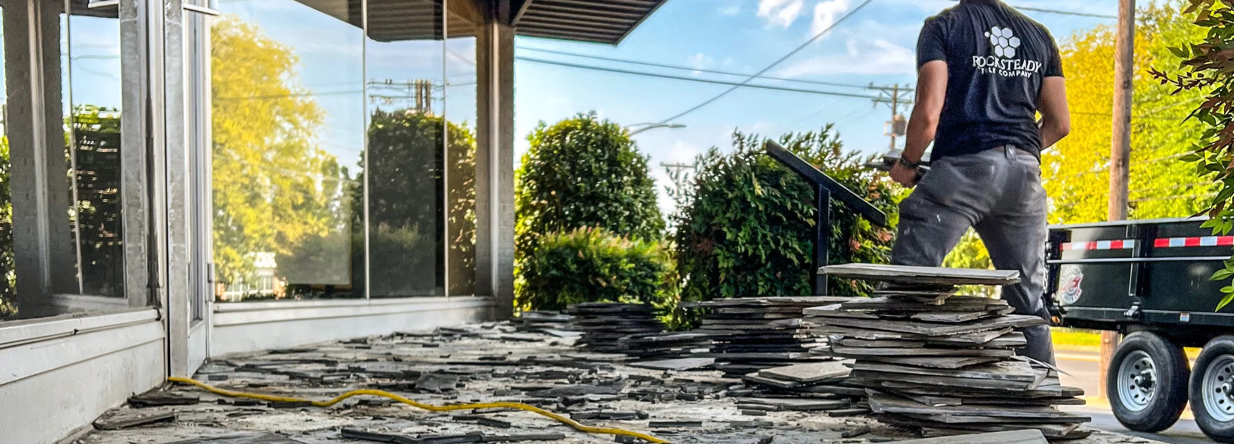 A person in a Rocksteady T-shirt discarding broken slate tiles on the porch of a building with glass windows, greenery, and a utility trailer in the background.