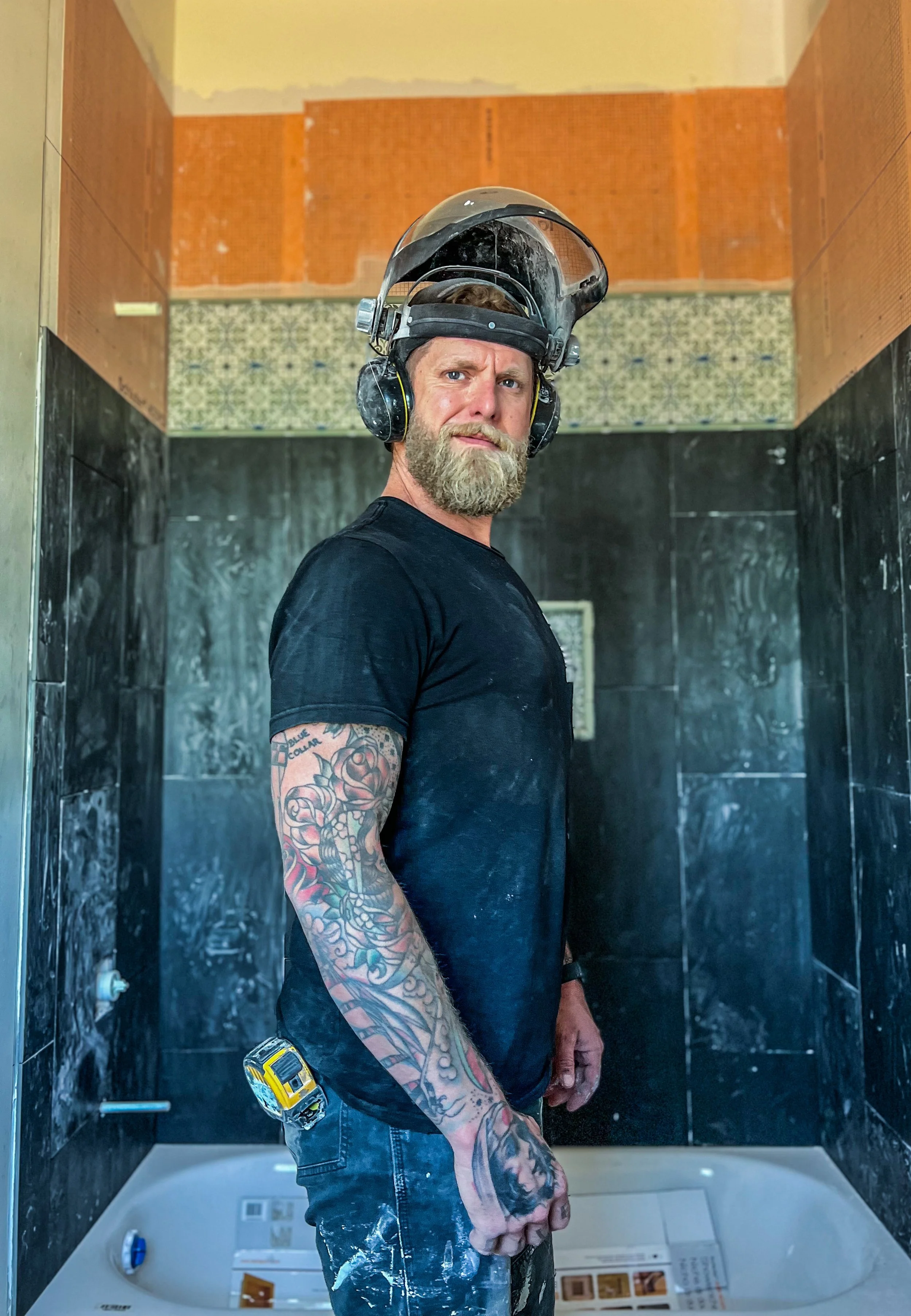 A tattooed man with a beard wearing a black t-shirt and a protective helmet stands inside a partially remodeled bathroom.