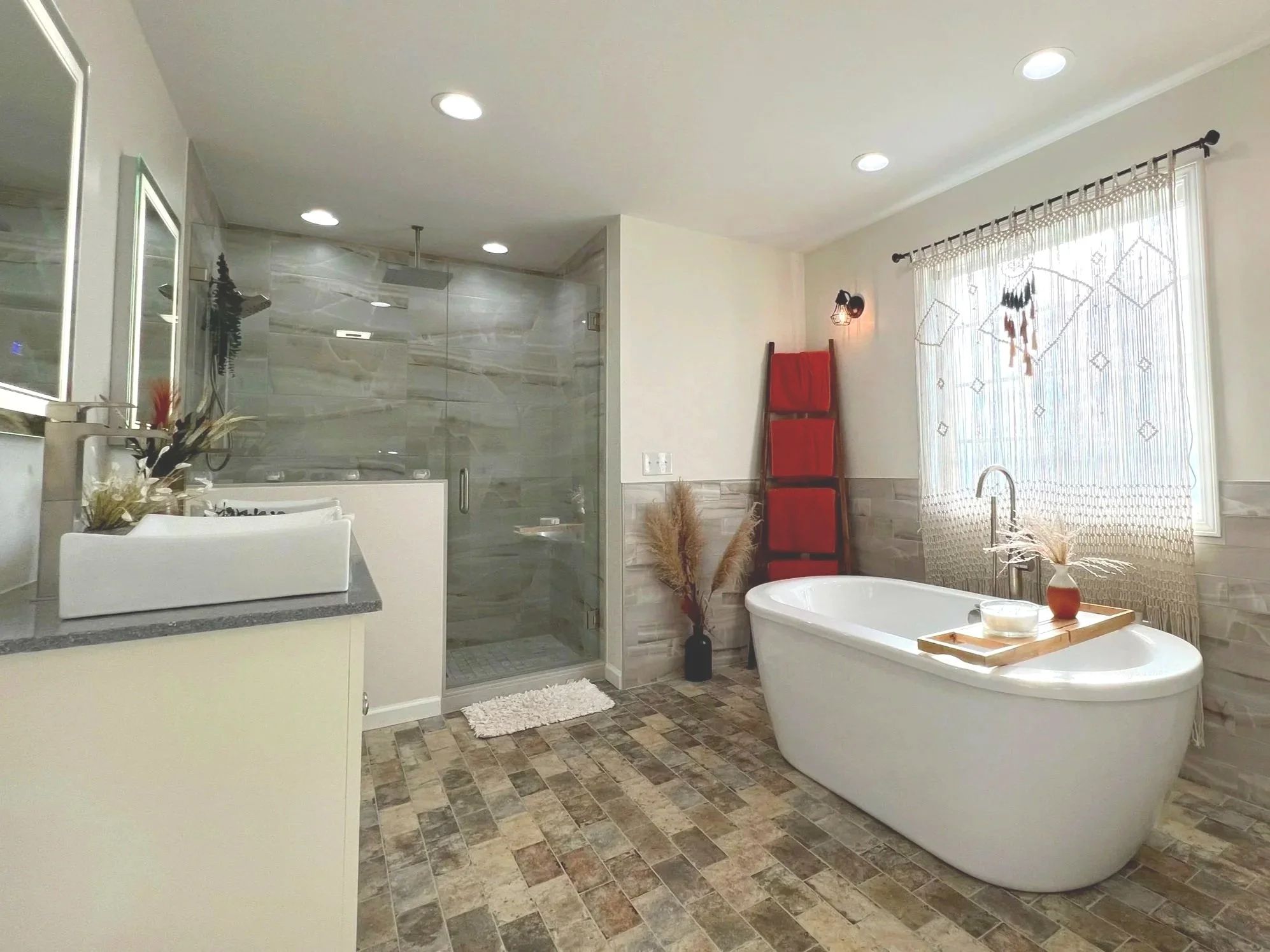 A modern bathroom with a freestanding bathtub near a window, a glass-enclosed shower area, a white vanity with a sink, and earthy tiles on the floor.