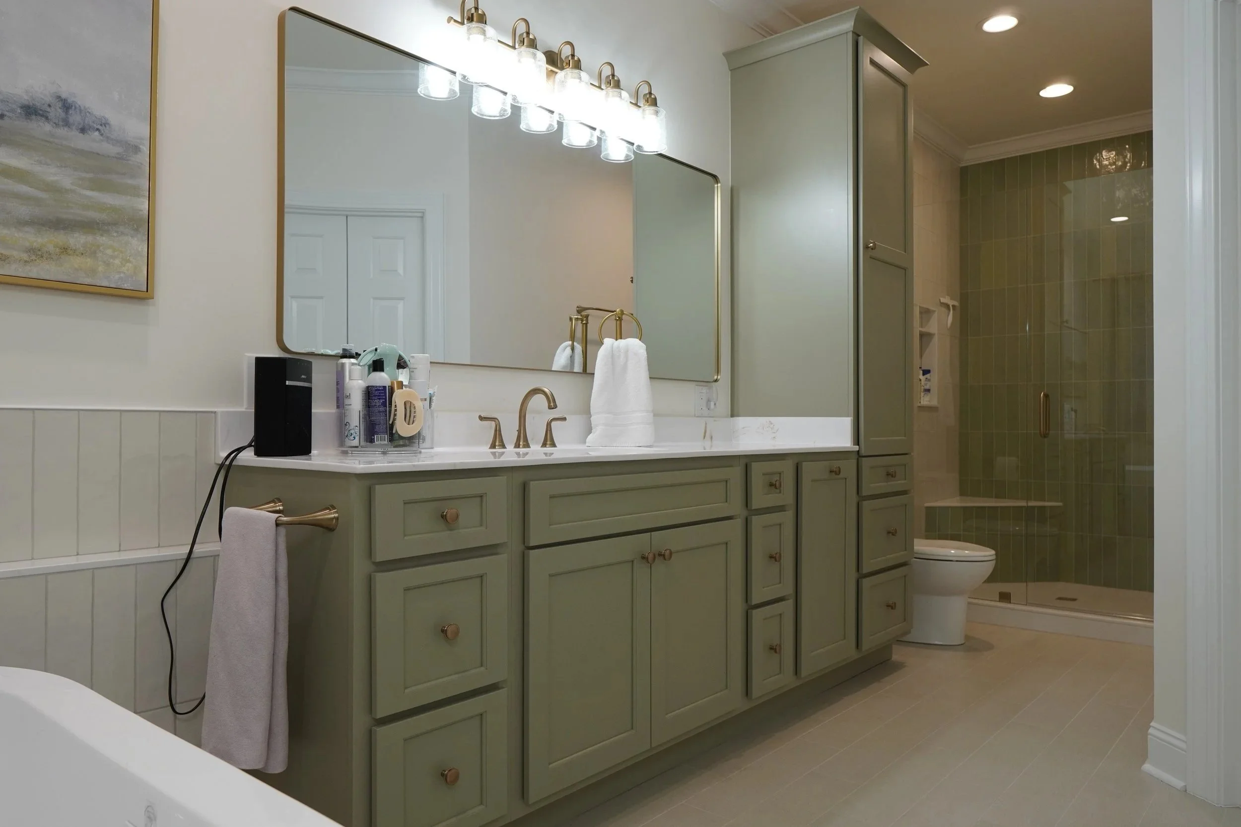 Green bathroom vanity with a large mirror, gold fixtures, and a variety of toiletries. Enclosed shower with green tiles and a glass door, toilet, and a corner cabinet for storage.