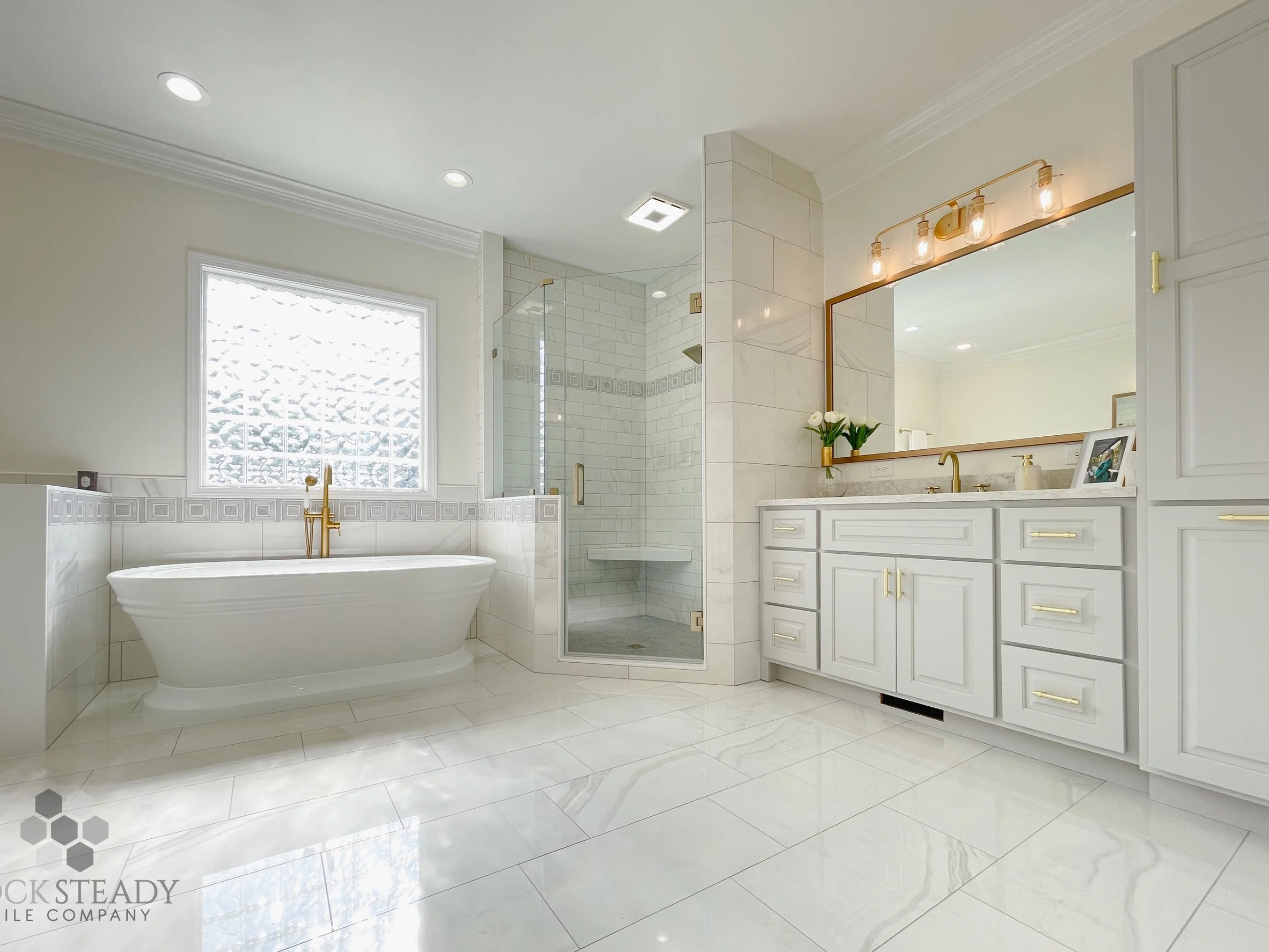 Bright, modern bathroom with white tile flooring, a large window, a freestanding bathtub, a glass-enclosed shower, and a white vanity with gold fixtures and a large mirror.