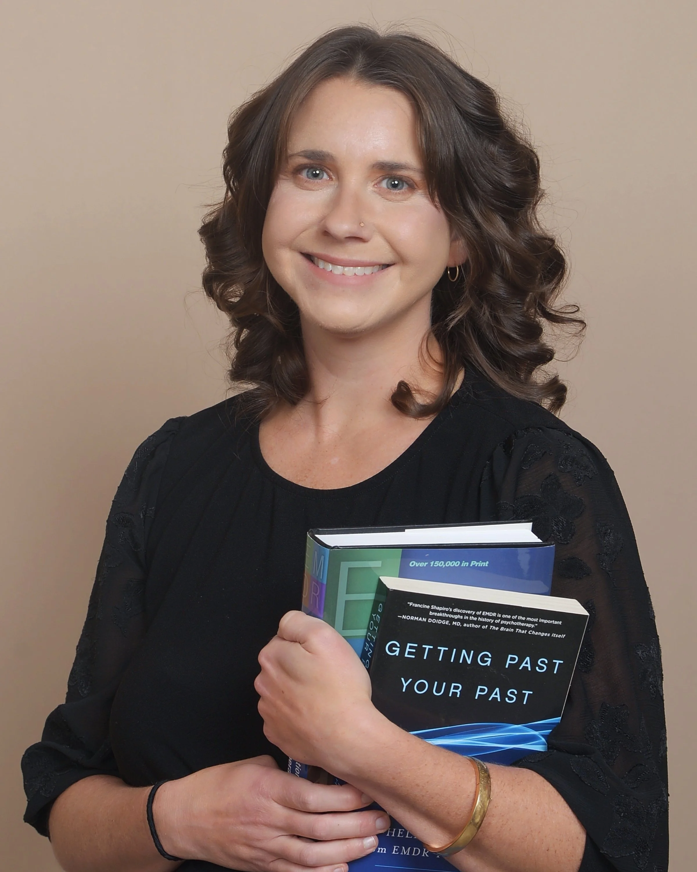 A woman smiling and holding a book titled "Getting Past Your Past." She has shoulder-length brown hair, wears a black shirt, a gold bracelet, a black hair tie on her wrist, a small nose piercing, and small hoop earrings. The background is beige.