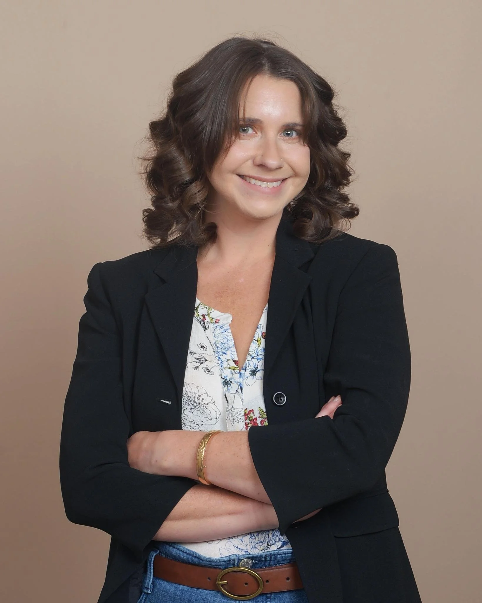 A woman with wavy brown hair and light skin smiling with arms crossed, dressed in a black blazer over a patterned blouse, wearing a bracelet, standing against a plain beige background.