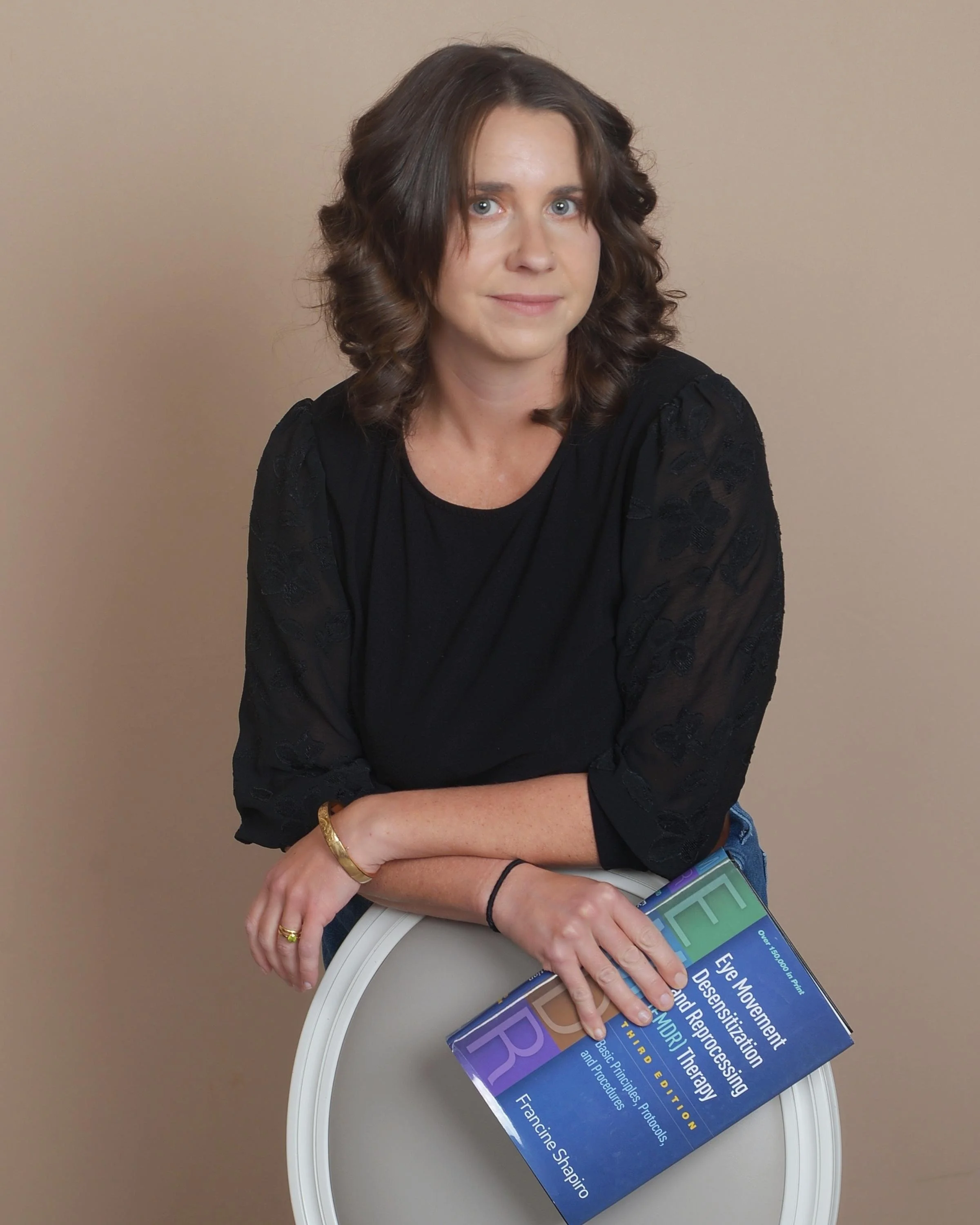 A woman with shoulder-length wavy brown hair and blue eyes seated at a white table, wearing a black top with lace sleeves, a gold bracelet, and a ring, holding a blue paperback book titled 'Eye Movement Desensitization and Reprocessing Therapy' by Francine Shapiro.