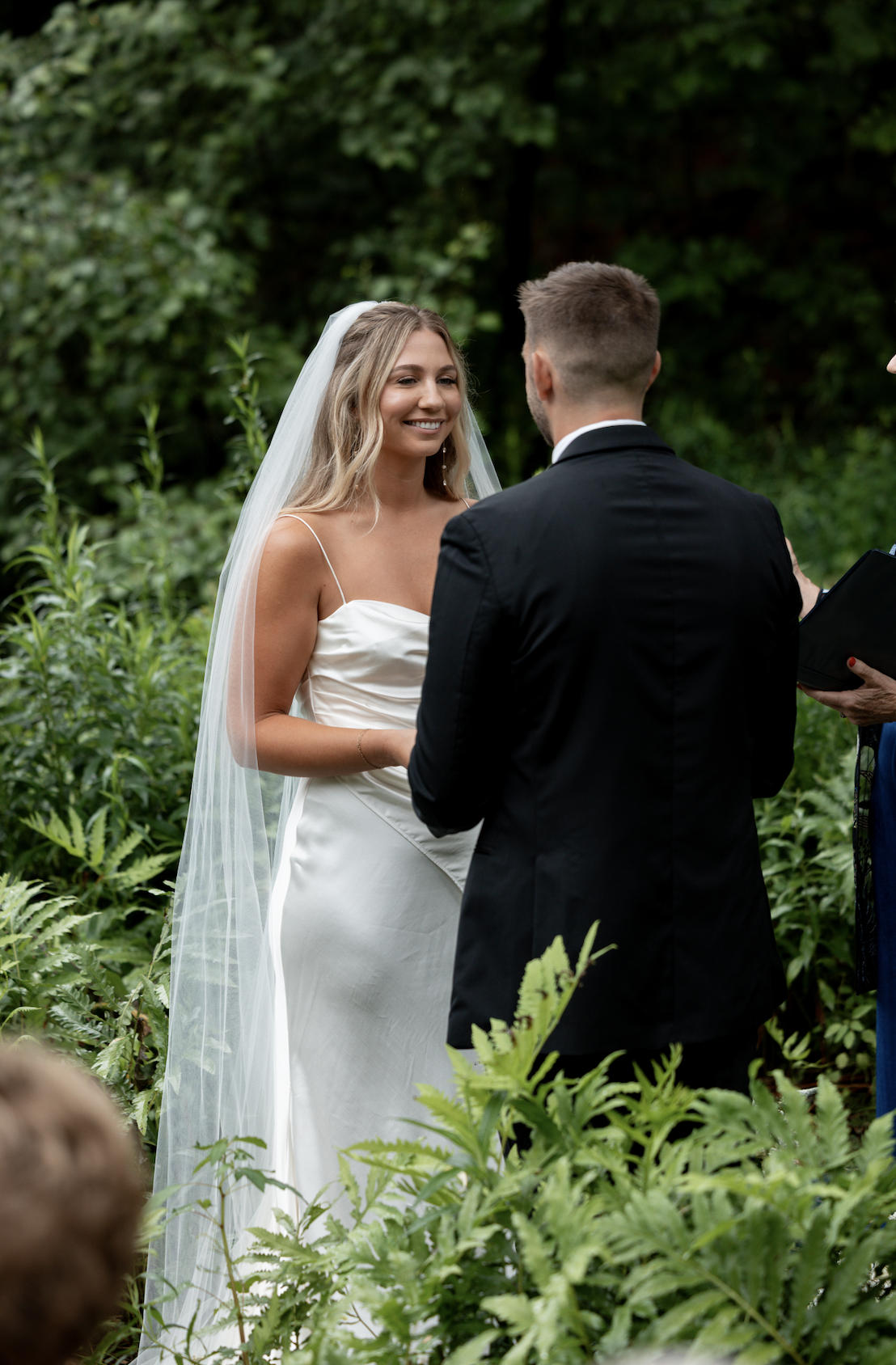 A bride and groom exchanging vows outdoors during a wedding ceremony surrounded by greenery.