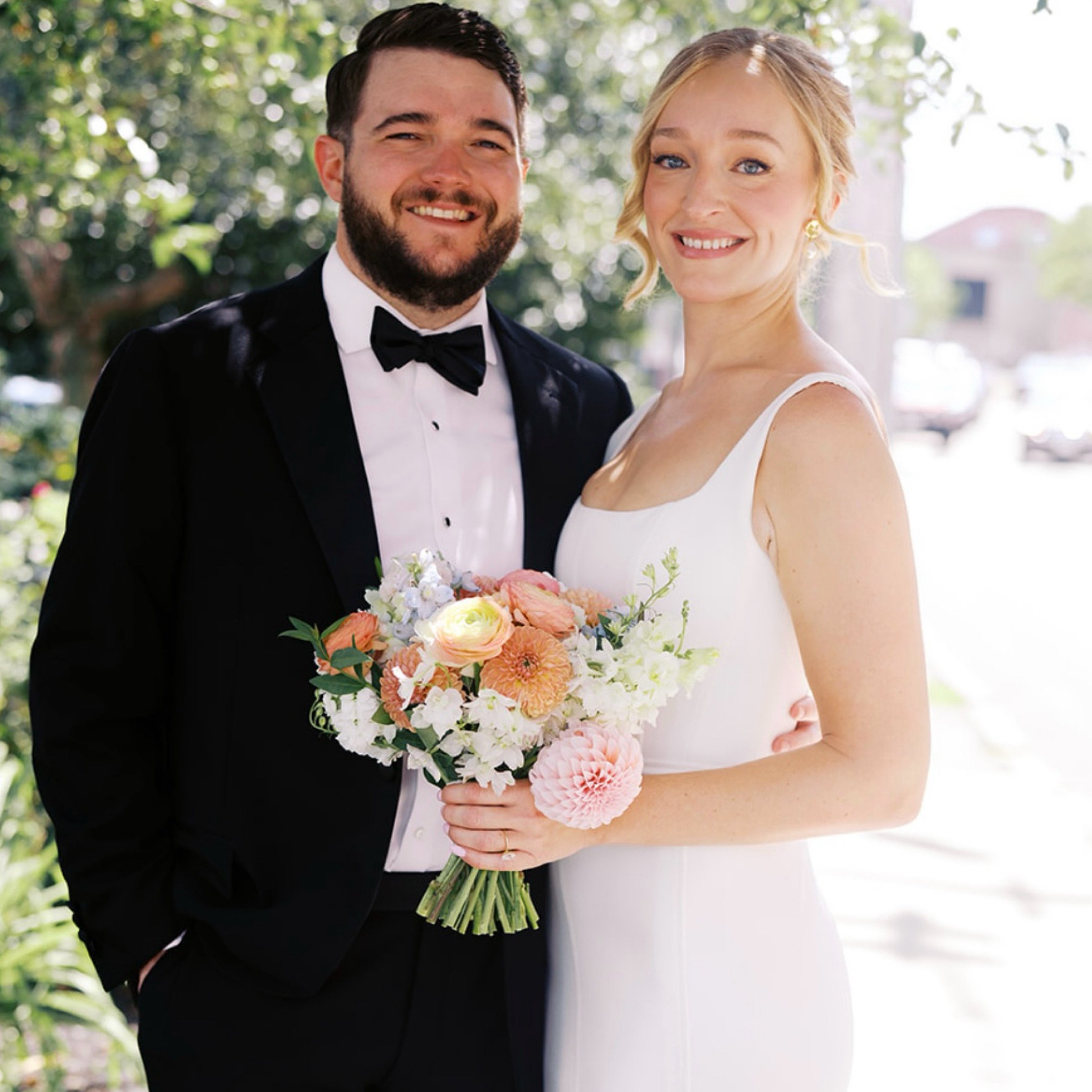 A newlywed couple smiling, the groom in a black tuxedo and bow tie, the bride in a white wedding dress holding a bouquet of pink, white, and peach flowers, standing outdoors on a sunny day.