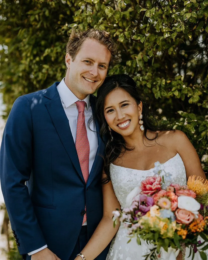 A newlywed couple smiling outdoors, with the man in a blue suit and pink tie, and the woman in a white wedding dress holding a bouquet of colorful flowers, standing near green shrubbery.
