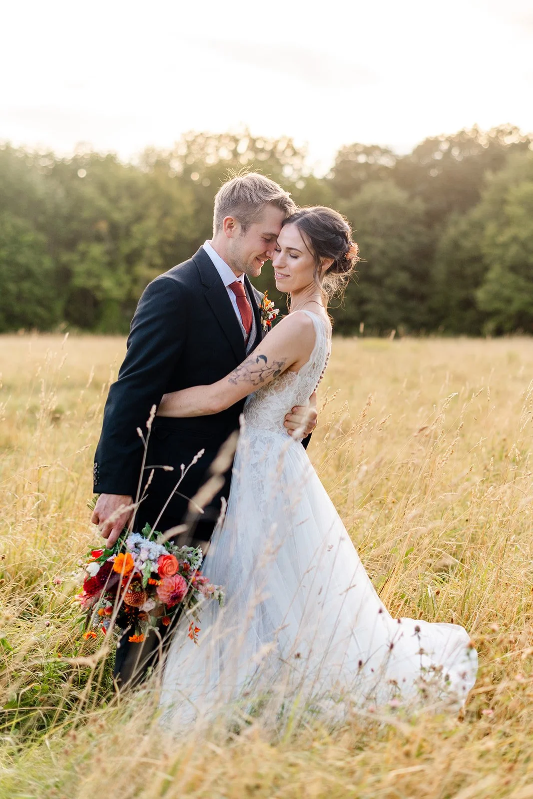 A newlywed couple embracing in a field of tall golden grass during sunset, with trees in the background. The groom is in a black suit holding a colorful bouquet, while the bride wears a white lace wedding dress with tattoos on her arm.
